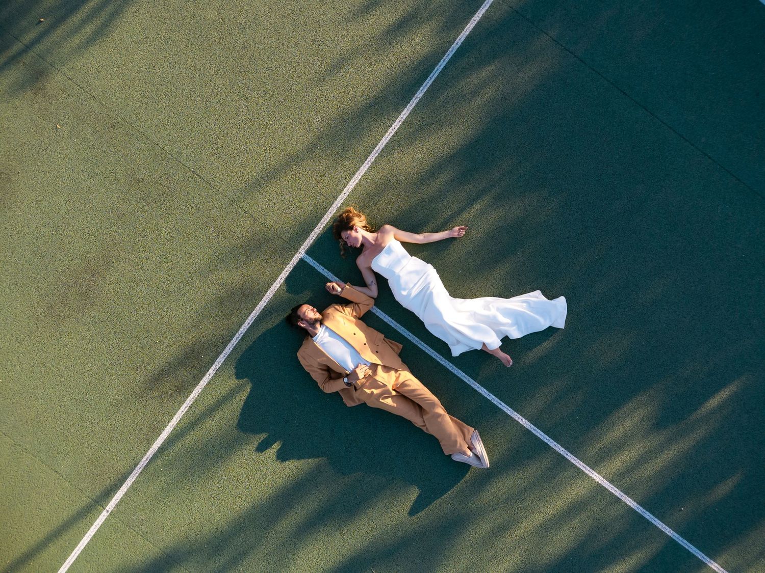 Aerial view of a bride and groom lying on a green tennis court in wedding attire. done wedding photographer