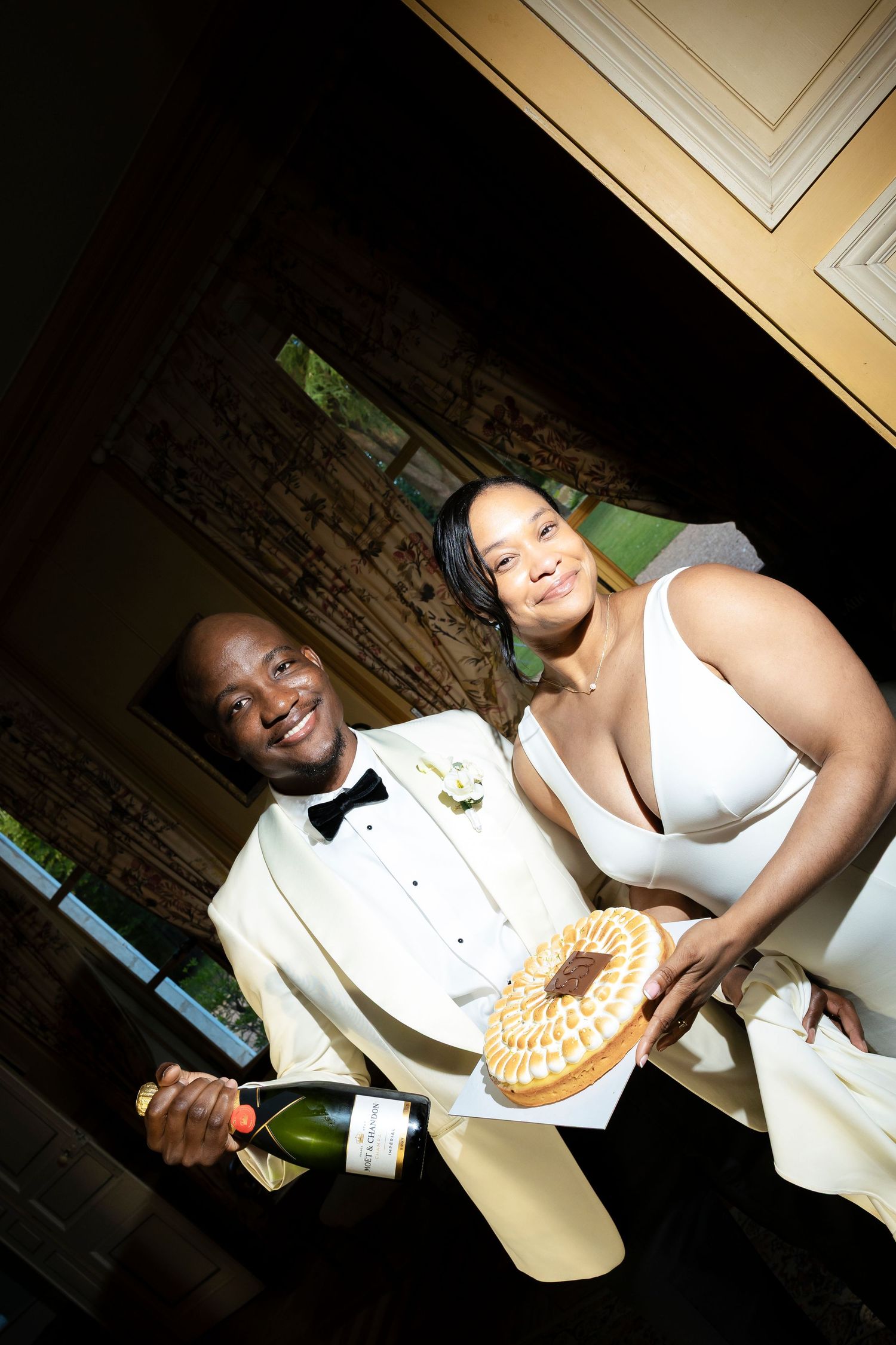 Smiling bride and groom pose with champagne and a decorative cake during their elegant wedding reception indoors.