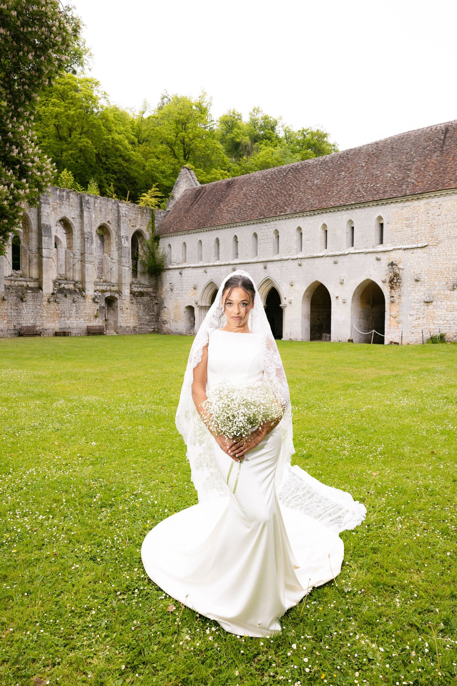 Bride in white mermaid gown with lace veil holding baby's breath bouquet outside historic abbey.