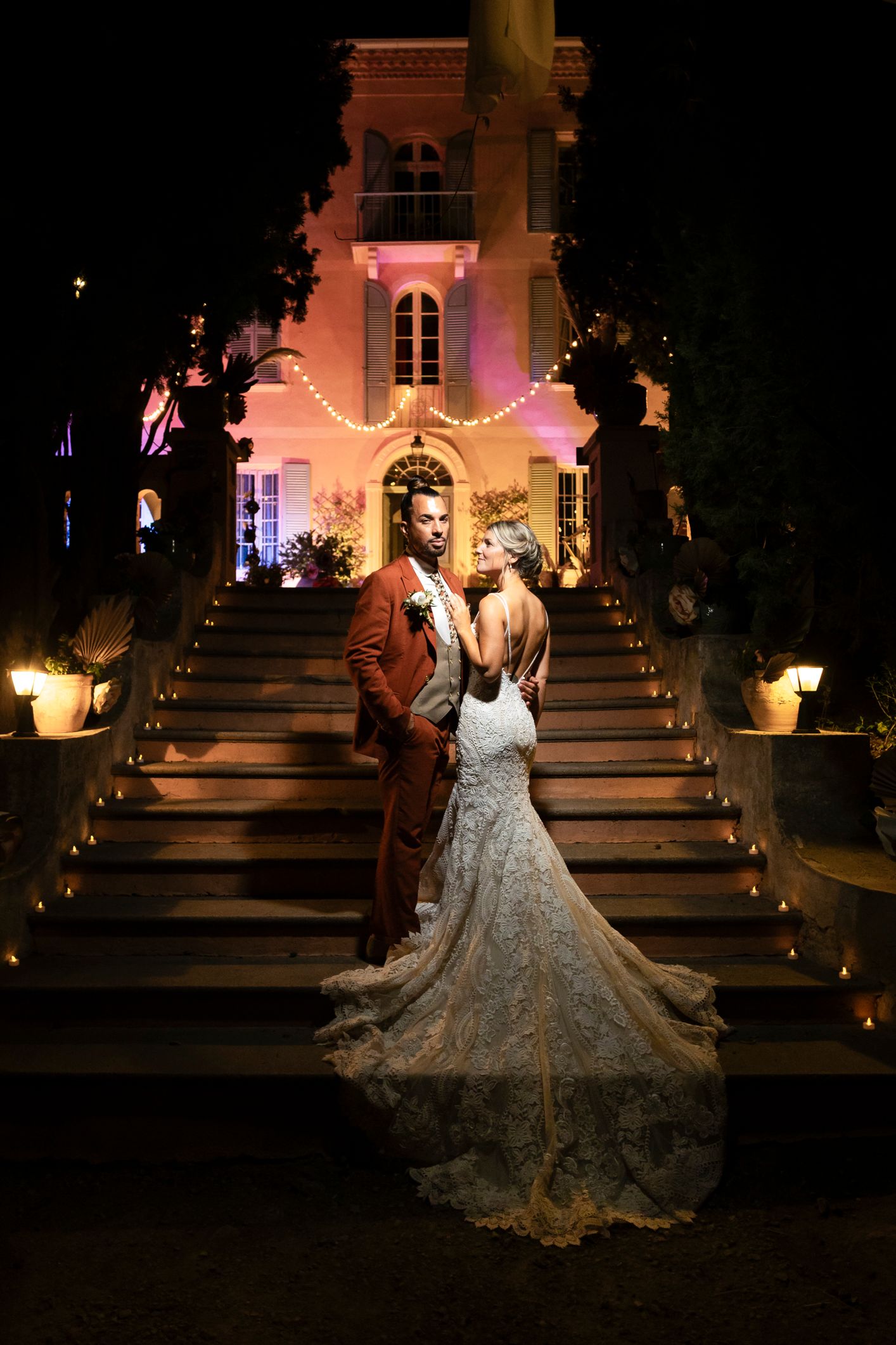 Bride and groom pose romantically on candlelit stairs of an elegant mansion glowing with pink lights at night.