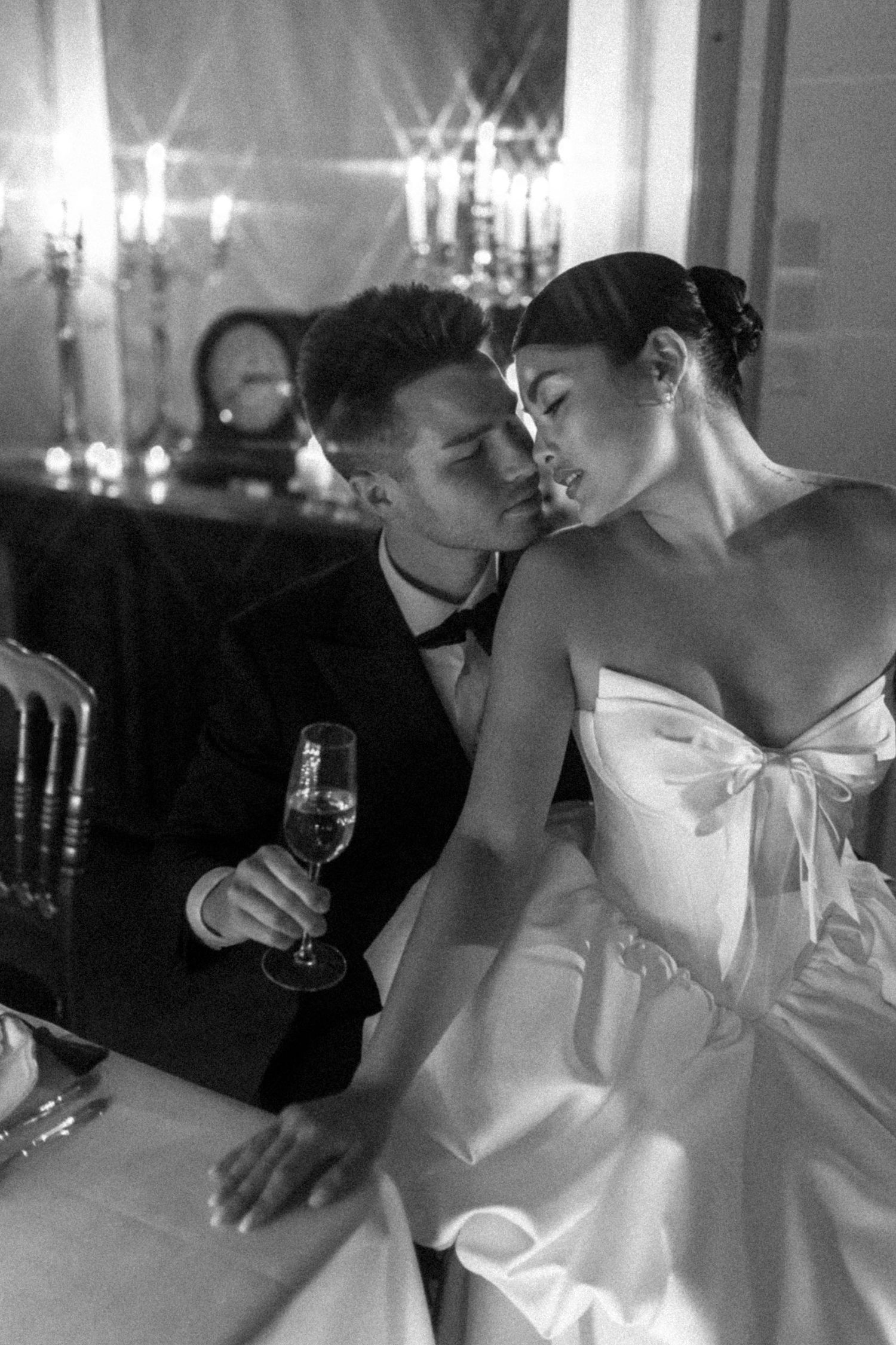 Black and white photo of a bride and groom sharing an intimate moment at a candlelit reception table, groom holding champagne.