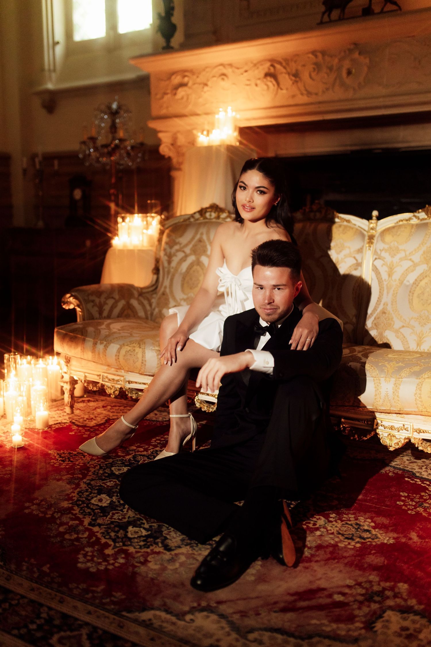 Elegant couple in formal attire posing in a luxurious candlelit room with ornate furniture and a grand fireplace.