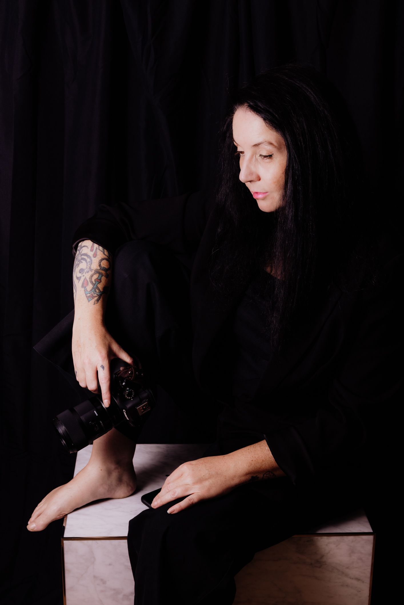 Woman in black outfit with tattoo sitting on white cube holding camera against dark background.