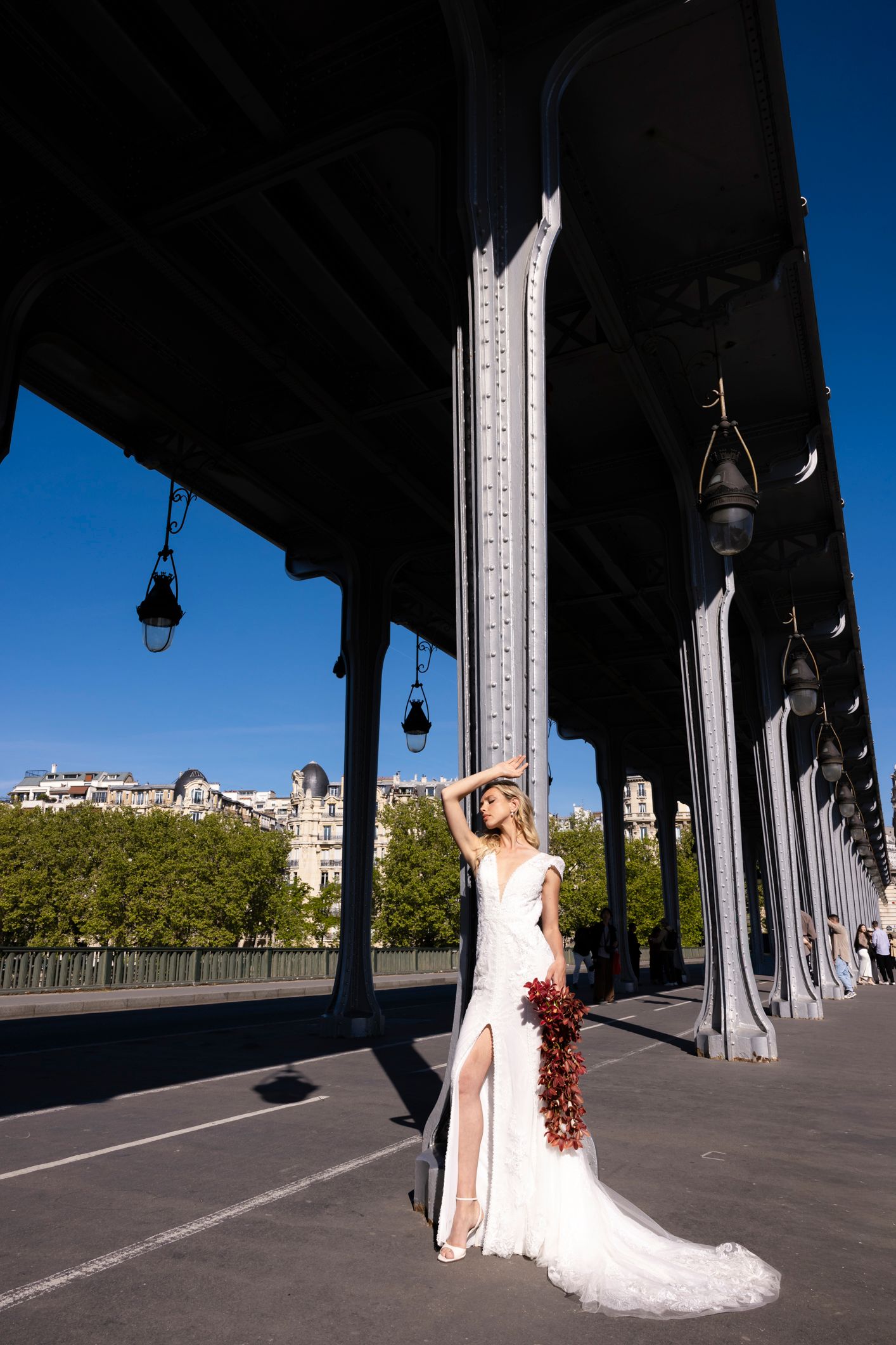 Bride in white gown holding bouquet under Bir-Hakeim Bridge, Paris, with blue sky and Haussmann buildings backdrop.