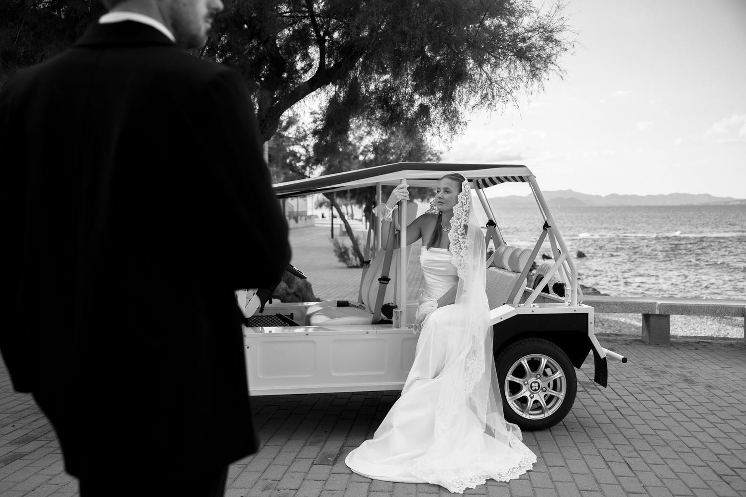 Black and white photo of a bride in a white gown stepping into a vintage golf cart by the ocean at a wedding.