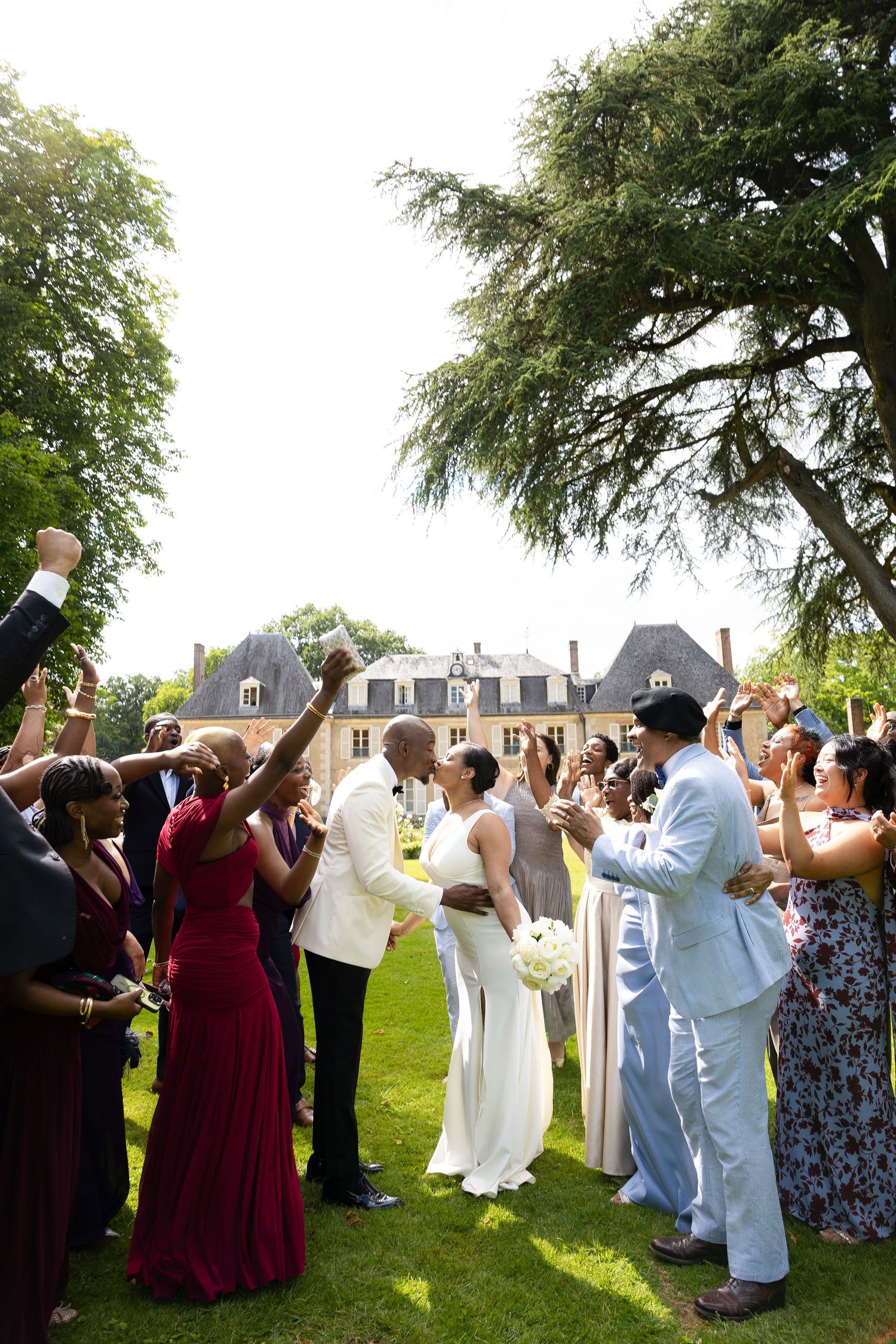 Joyful newlyweds share a kiss surrounded by celebrating guests on a sunny lawn outside an elegant French château.