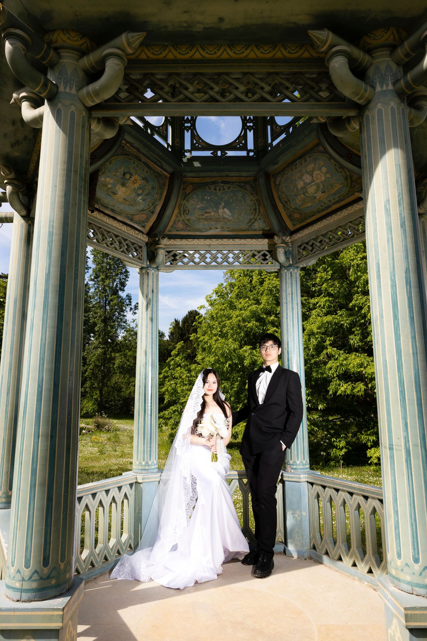 Bride and groom pose in elegant ornate gazebo with classical columns surrounded by lush green trees on their wedding day.