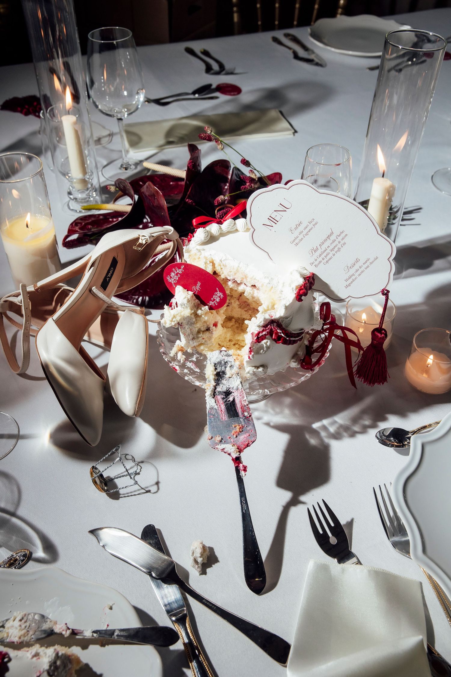Elegant wedding table with white cake, candles, heels, and red ribbon decor on white linen.