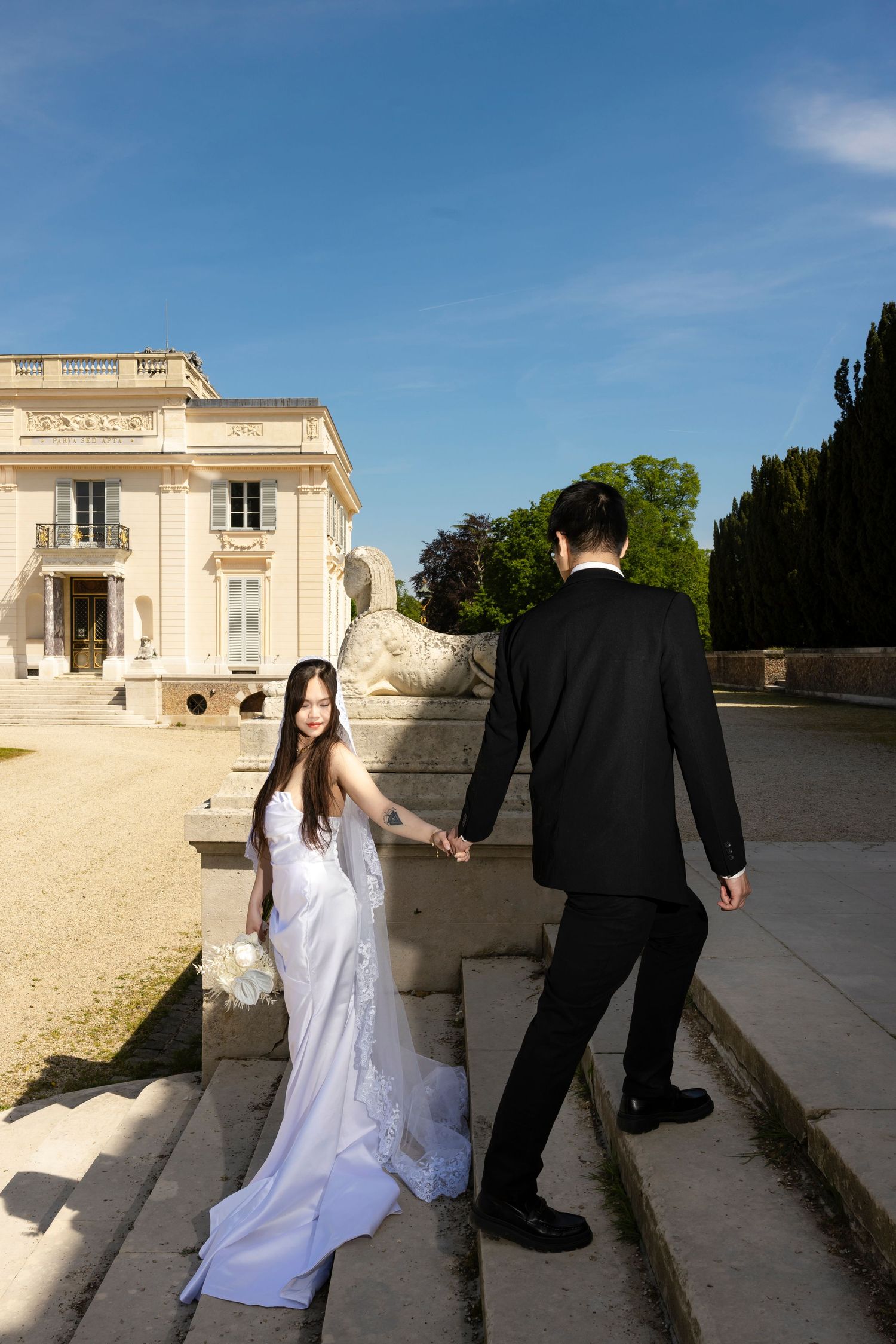 Bride and groom holding hands on stone steps outside elegant French chateau on sunny wedding day.