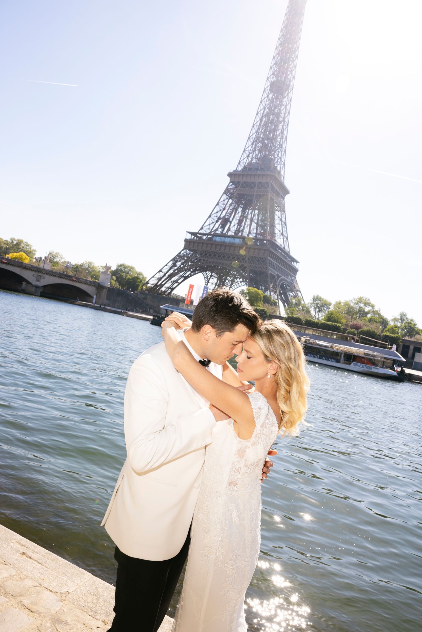 photographe mariage paris Couple embracing romantically in front of the Eiffel Tower by the Seine River in Paris on a sunny day.