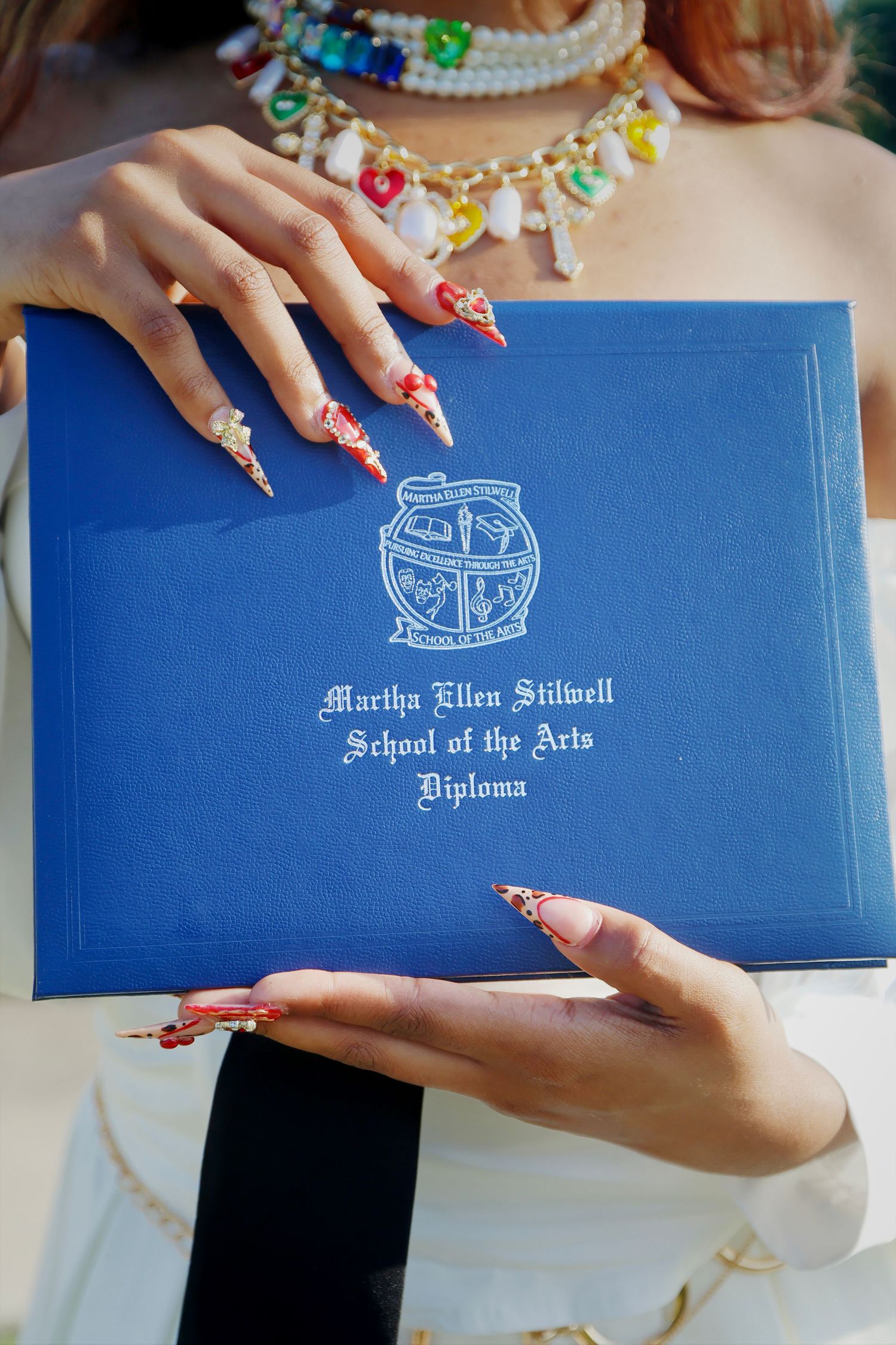 Hands with long red decorated nails holding a blue Martha Ellen Stilwell School of the Arts diploma.