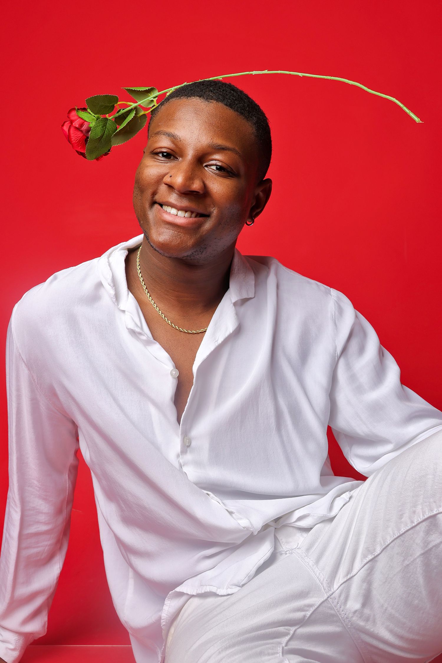 Smiling young man in white shirt and pants holding a red rose against a vibrant red background.