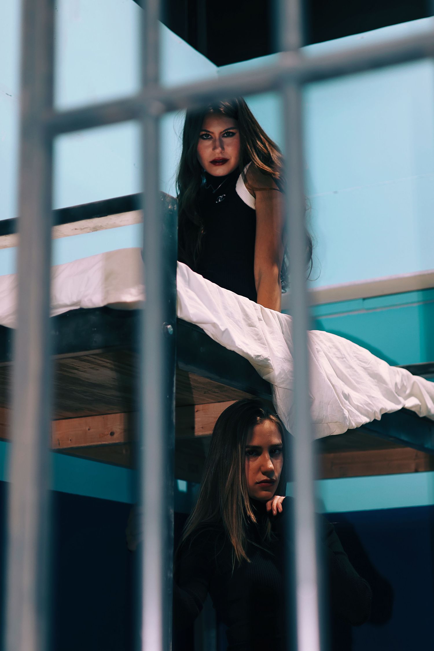 Two women in black outfits pose dramatically behind bars, one on a bunk bed above, in a moody blue-toned setting.