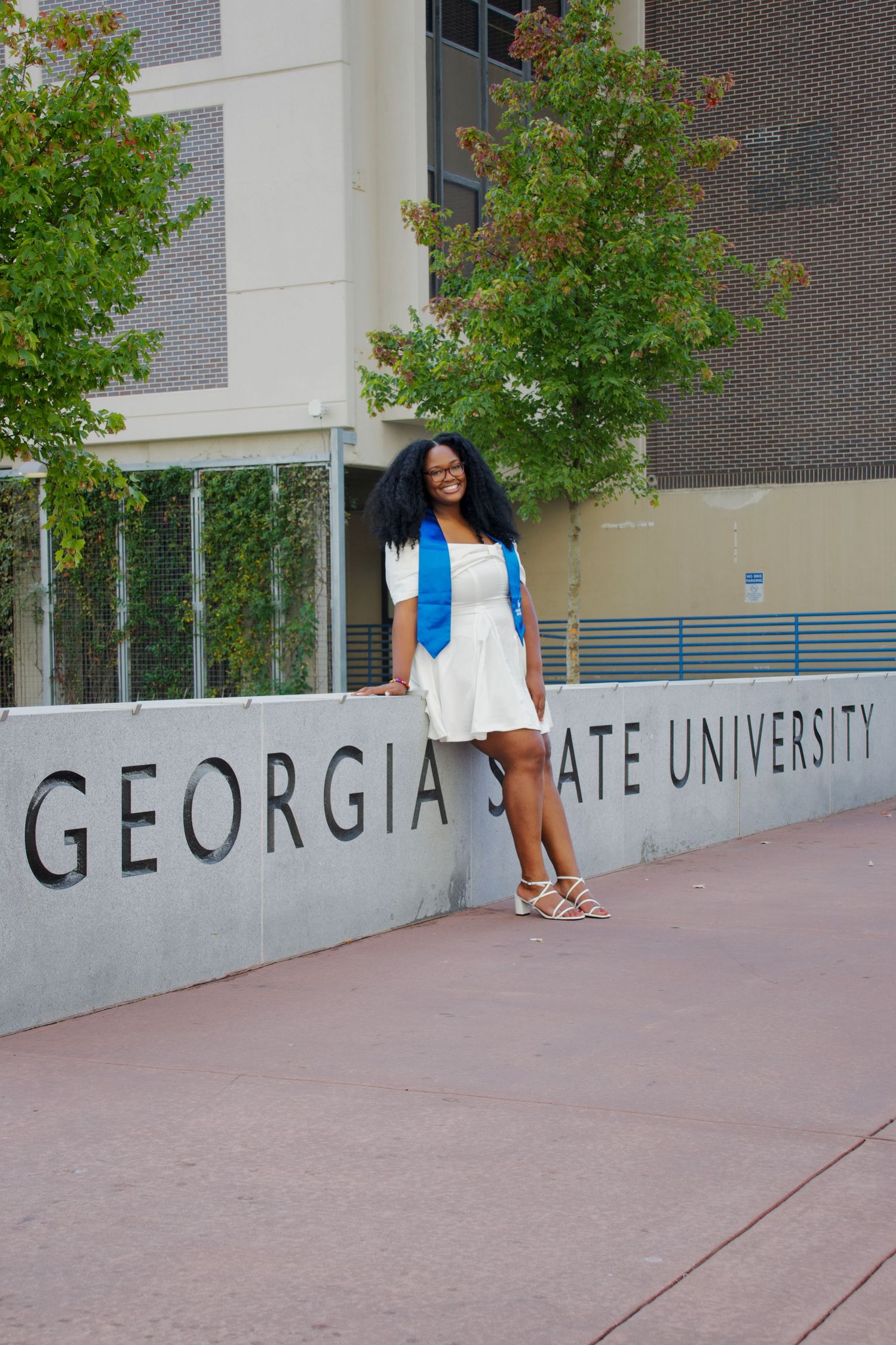 Smiling female graduate in white dress with blue stole poses at Georgia State University sign.
