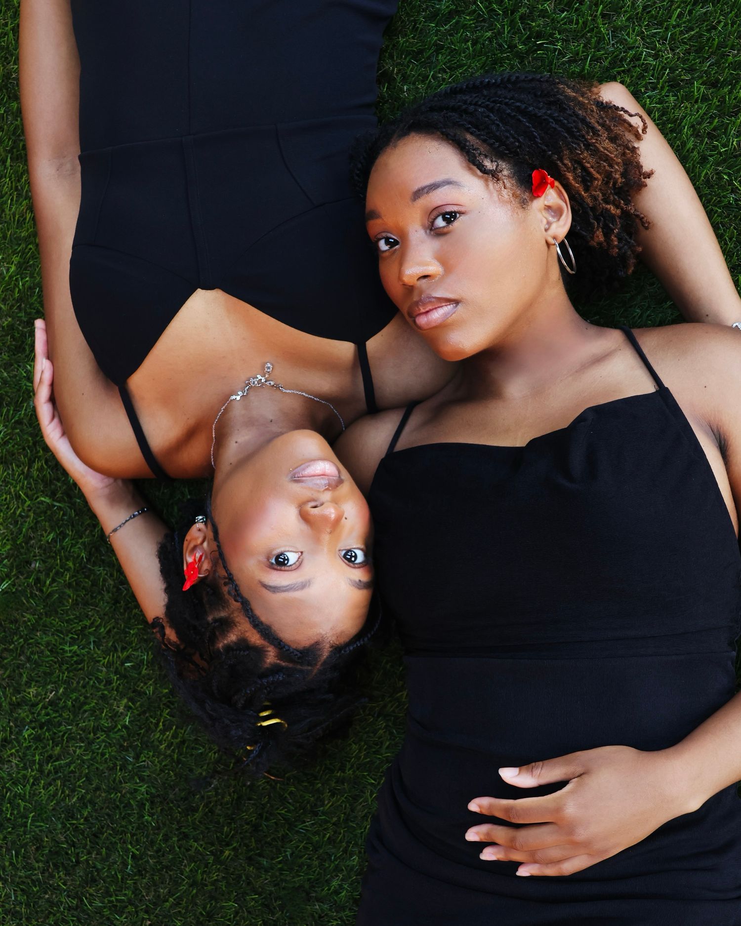 Two young Black women in black outfits lying on green grass, looking up at camera in a creative portrait.