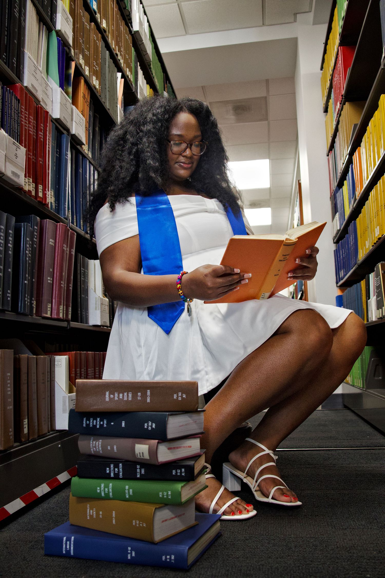 Female graduate in white dress and blue stole reads a book while sitting in a library.