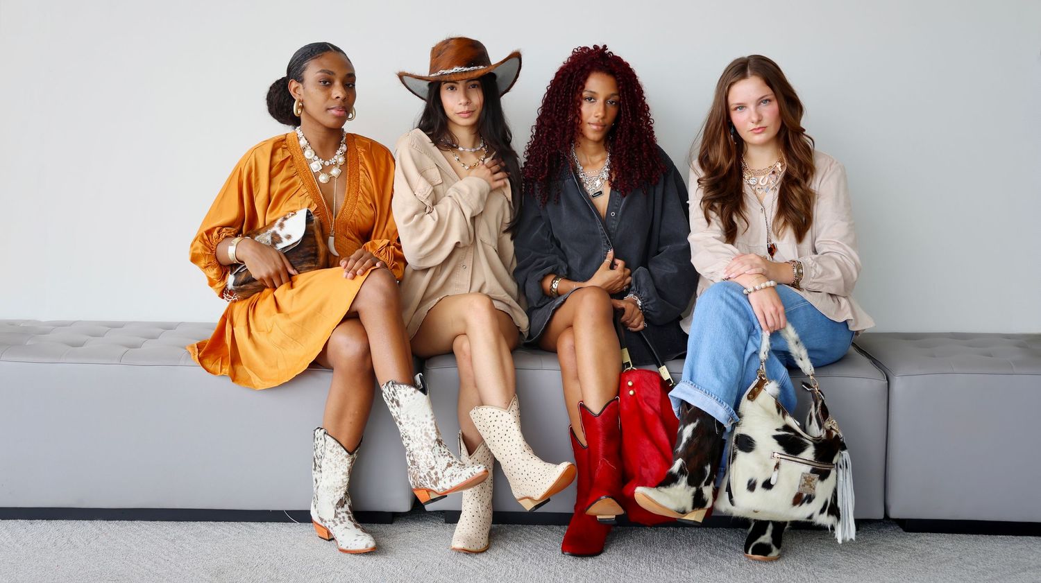 Four stylish women in colorful western-inspired outfits with boots and bags seated against white wall.