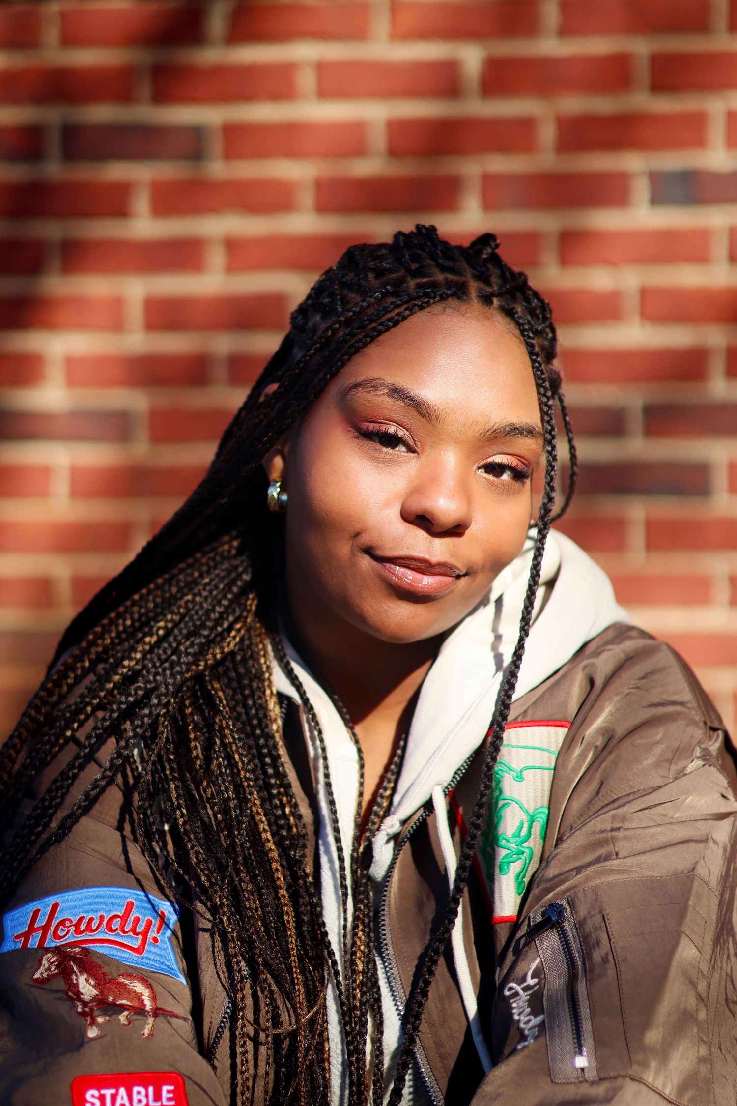 Young Black woman with long box braids wearing a patch jacket poses against a sunny brick wall.