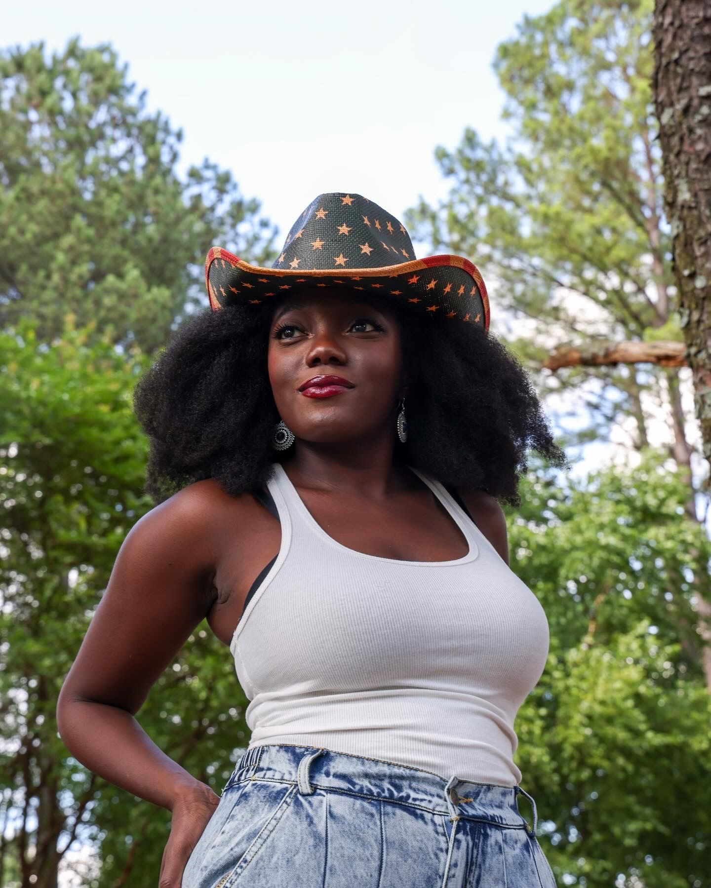 A Black woman in a white tank top, jeans, and a star-patterned cowboy hat poses outdoors among tall green trees.