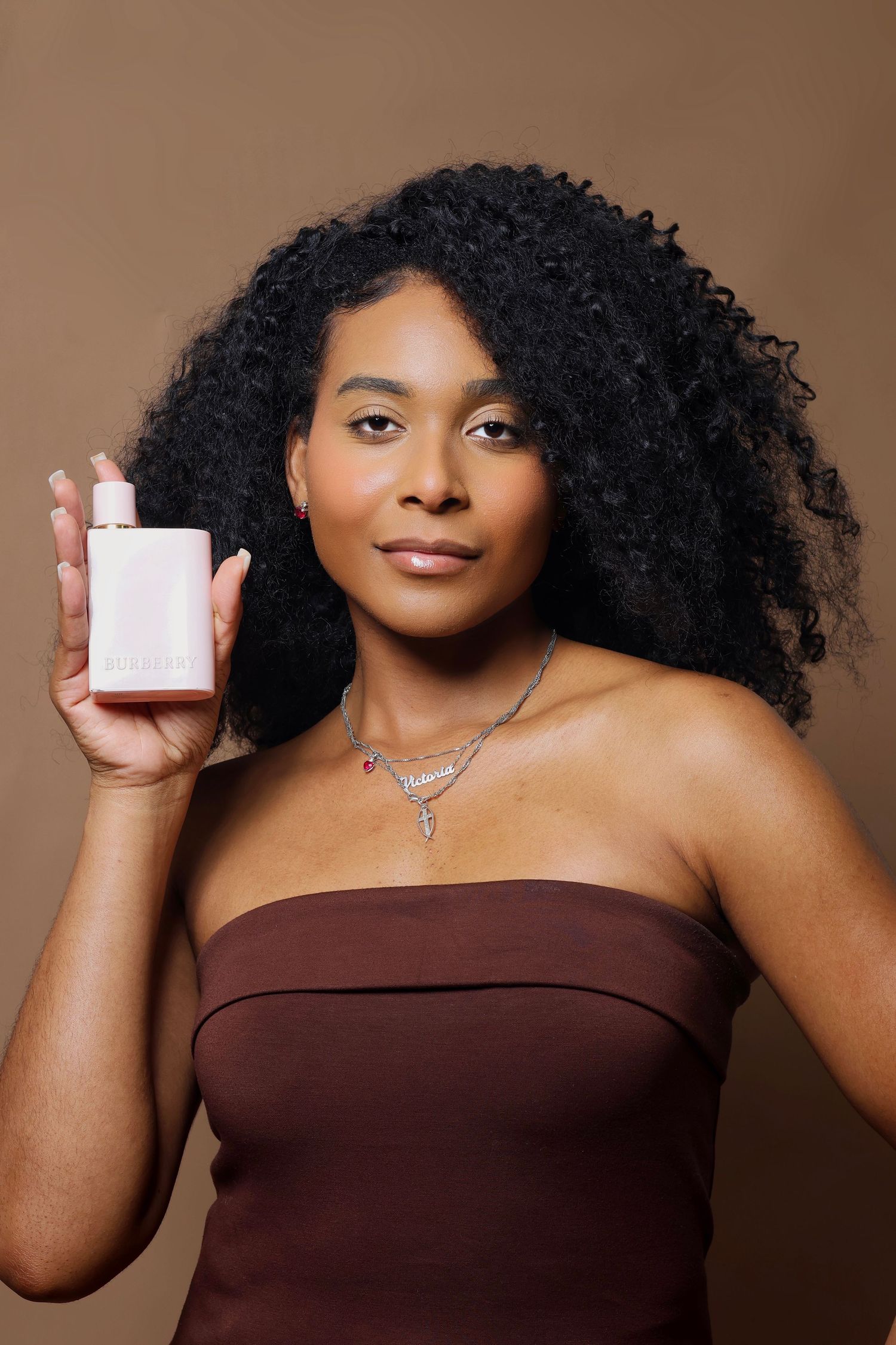 Woman with curly hair holding a white fragrance bottle wearing a brown strapless top and layered necklaces.