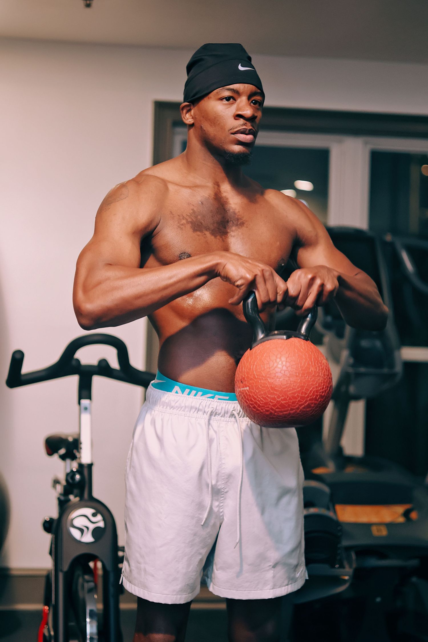 Muscular shirtless Black man holding a kettlebell in a gym, wearing white Nike shorts and a black cap.