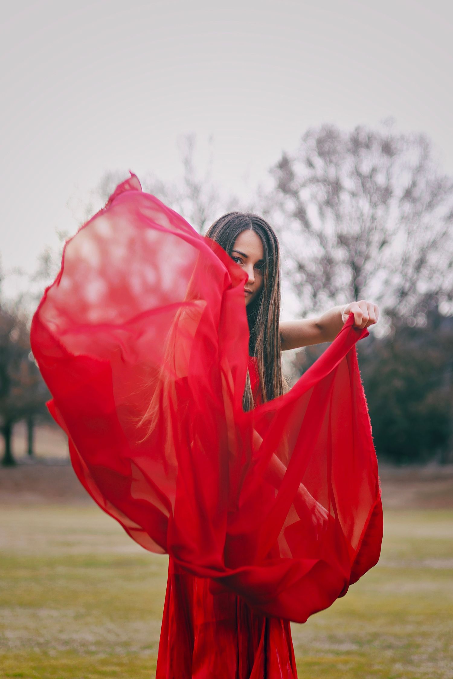 Woman in red dress holding flowing red fabric in outdoor park setting.