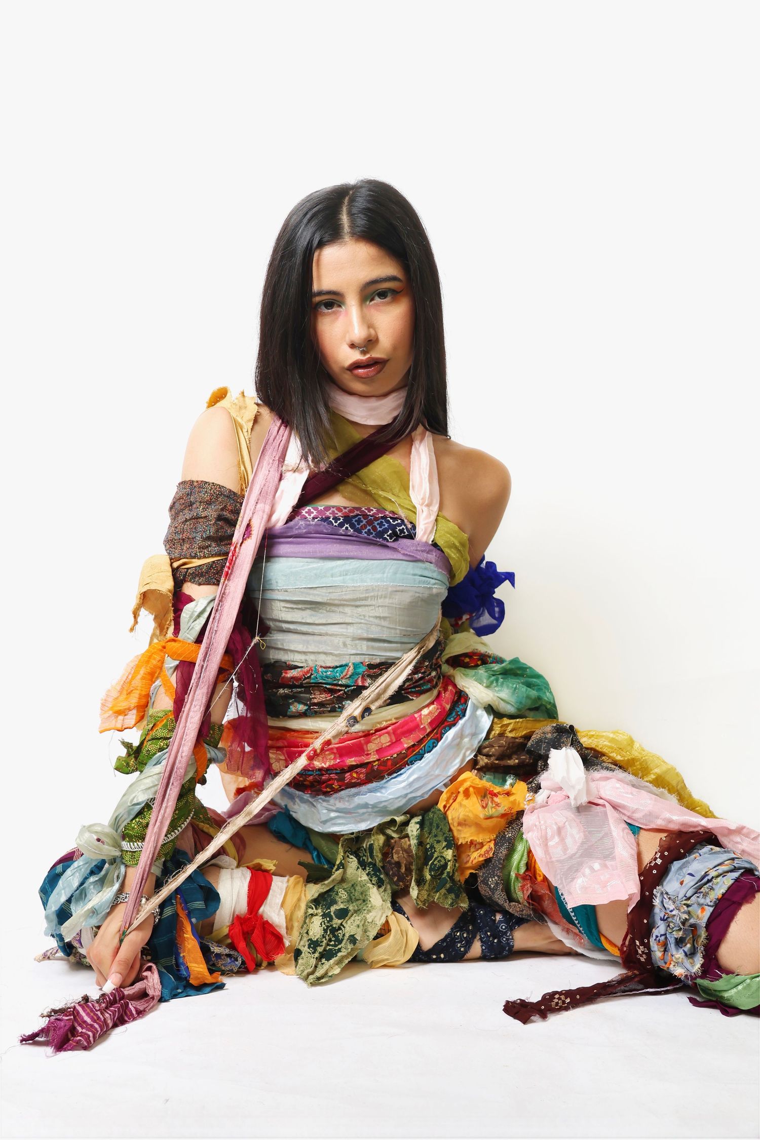 Young woman sitting on floor wrapped in colorful fabric scraps and textile pieces in artistic fashion editorial.