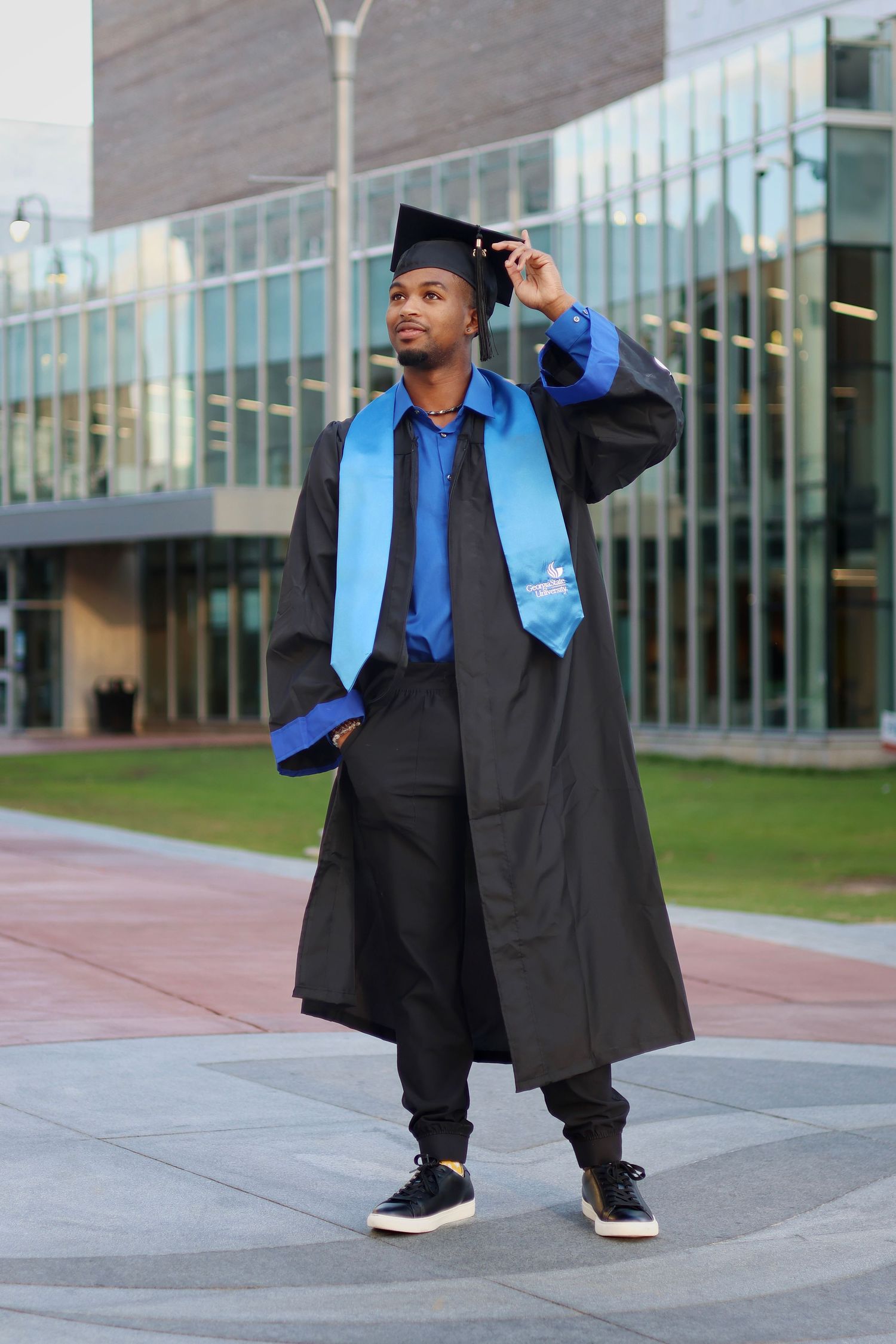 Male graduate in black and blue cap and gown poses confidently outside a modern building.