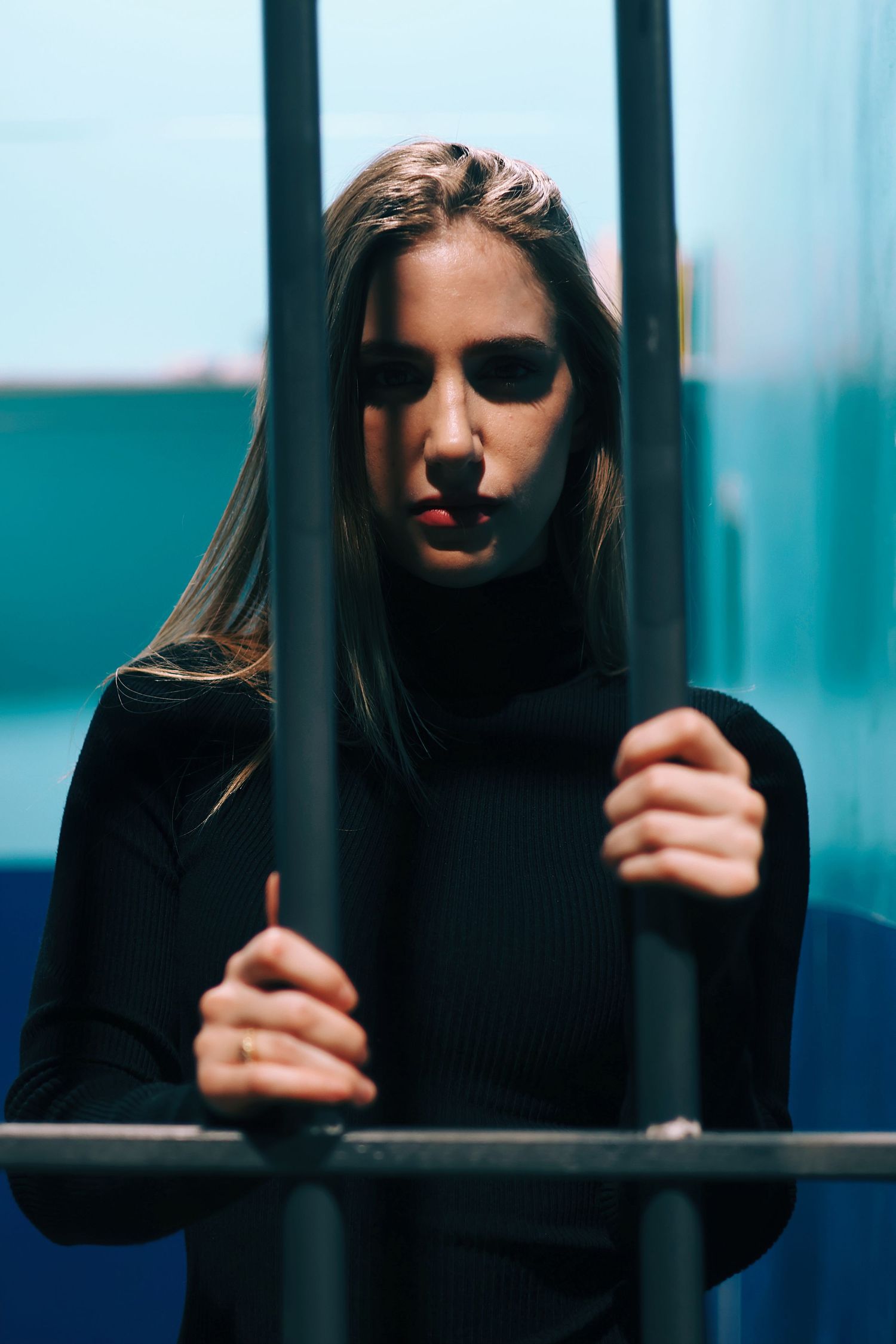 Young woman with long hair gripping metal prison bars, looking down with a somber expression in a jail cell.