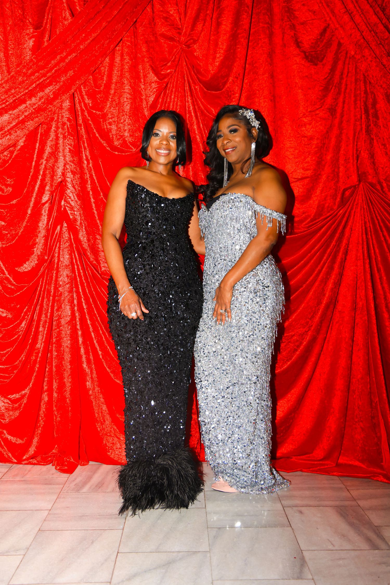 Two women in elegant sequined gowns pose in front of a red draped backdrop at a formal event.