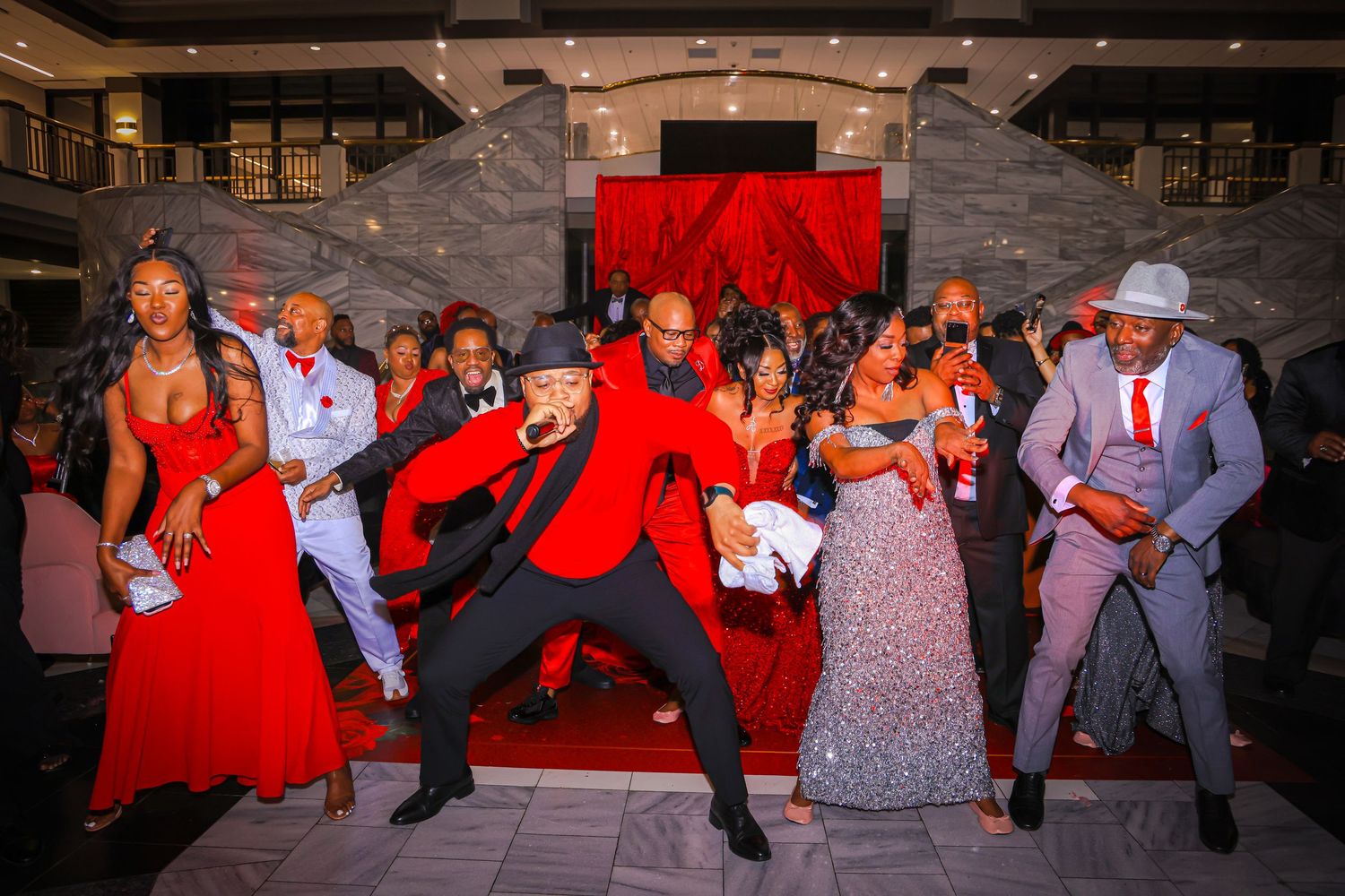 Guests in red and gray formal attire dance energetically at an elegant event with red carpet decor.