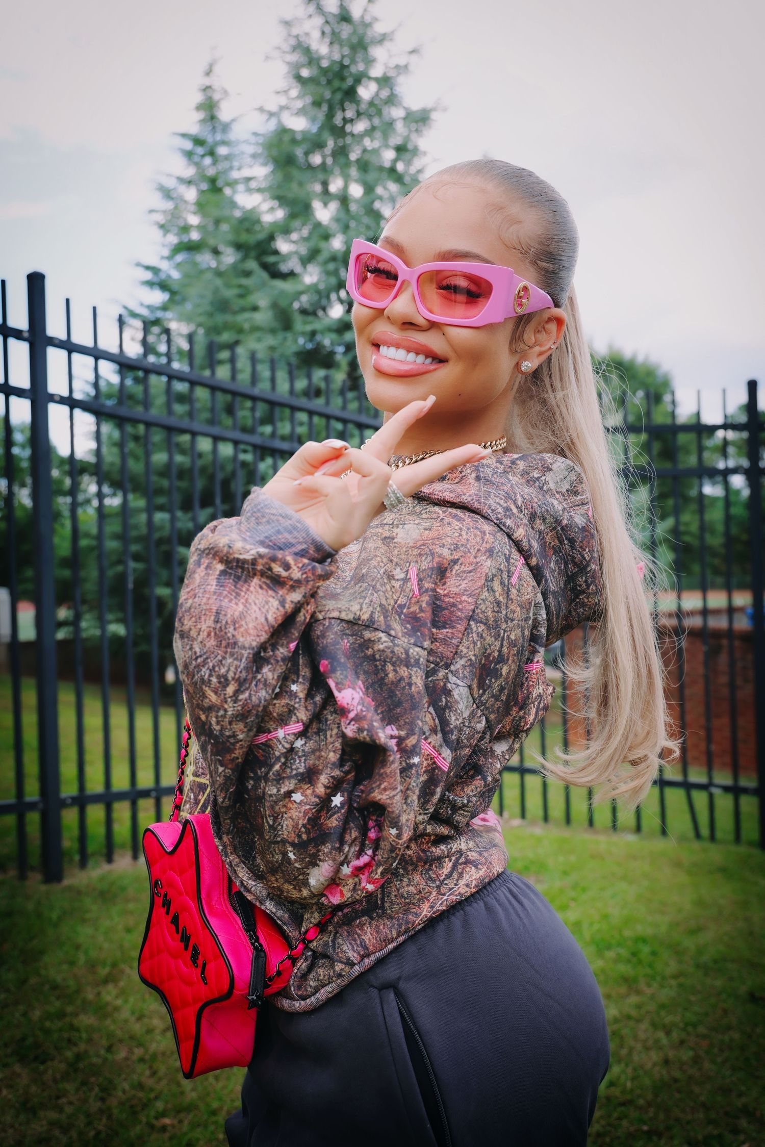 Smiling woman wearing pink sunglasses and floral top holding pink bag outdoors near black iron fence.