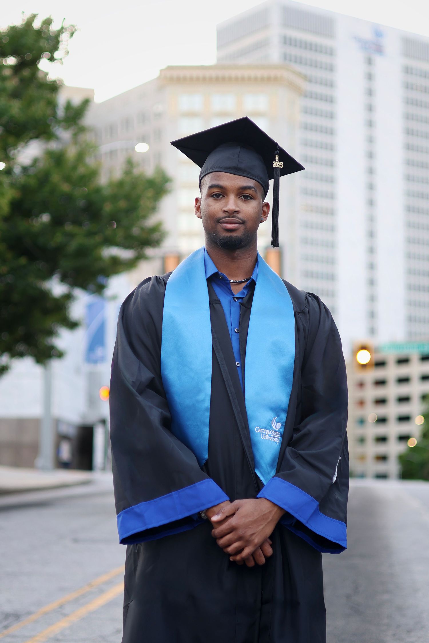 Young Black male graduate in black cap and gown with blue stole stands proudly on urban street with city buildings behind him.