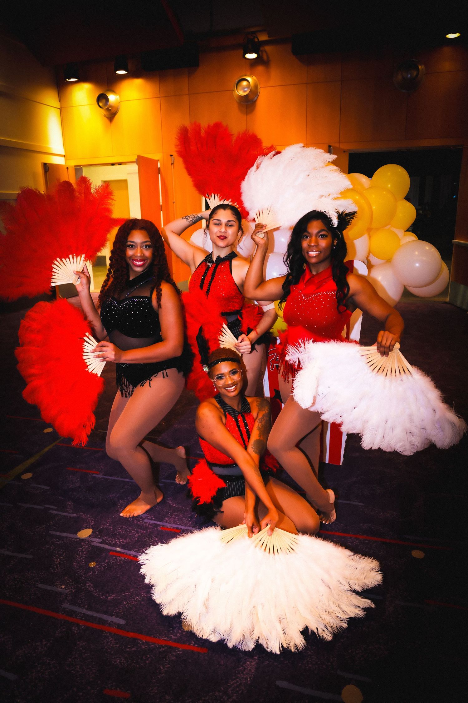 Four dancers in red costumes pose with large feather fans at a vibrant performance event.