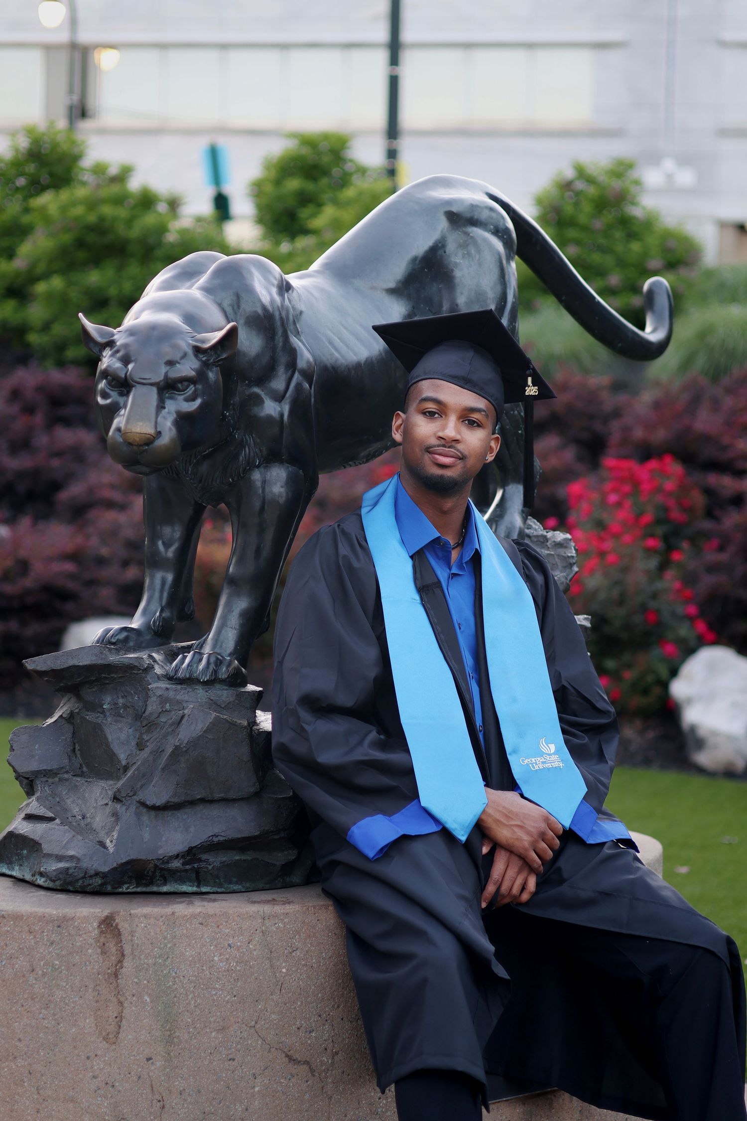 Graduate in cap and gown poses beside a panther statue on a college campus.
