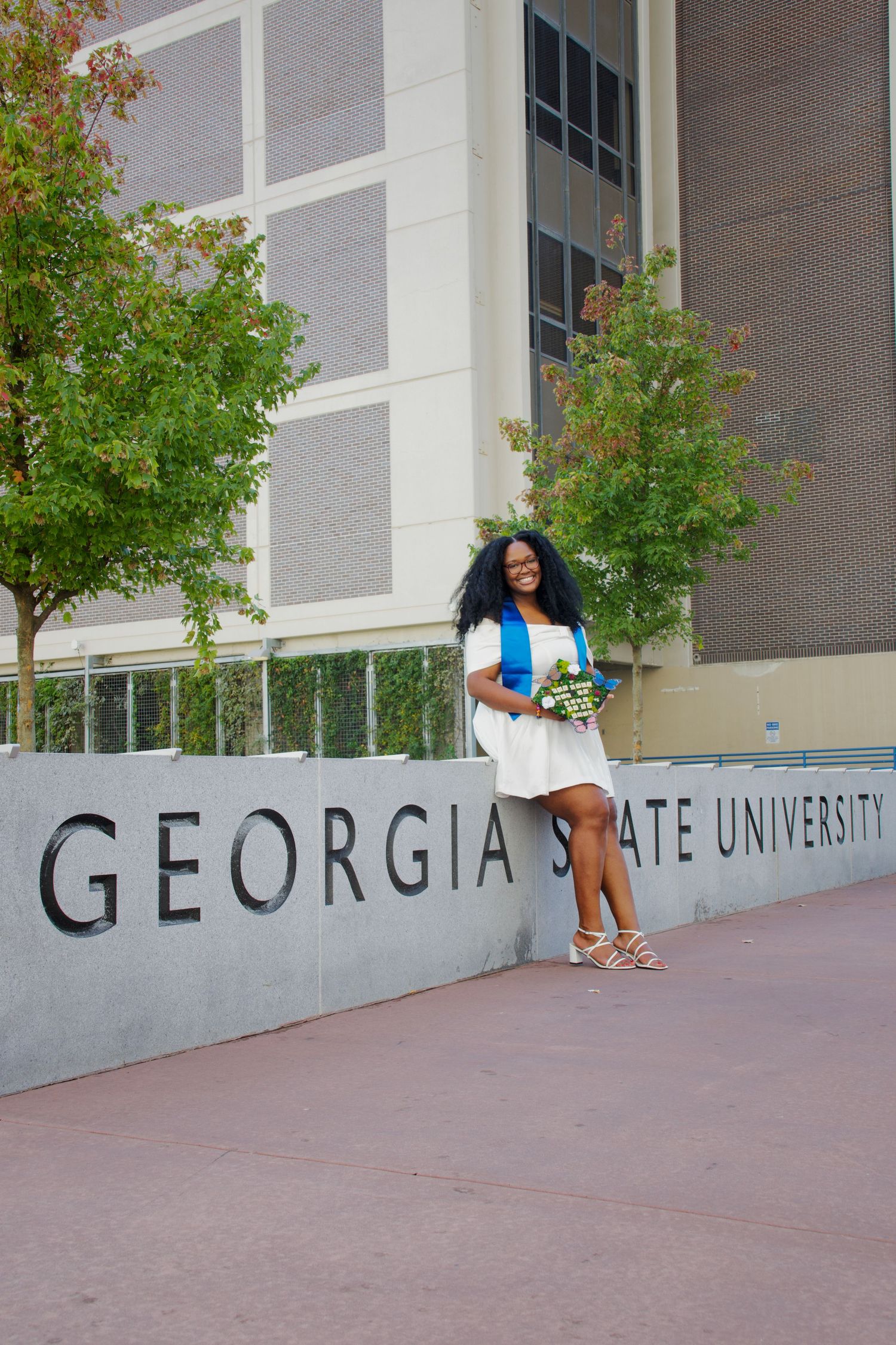 Smiling graduate in white dress with blue sash posing at Georgia State University sign.