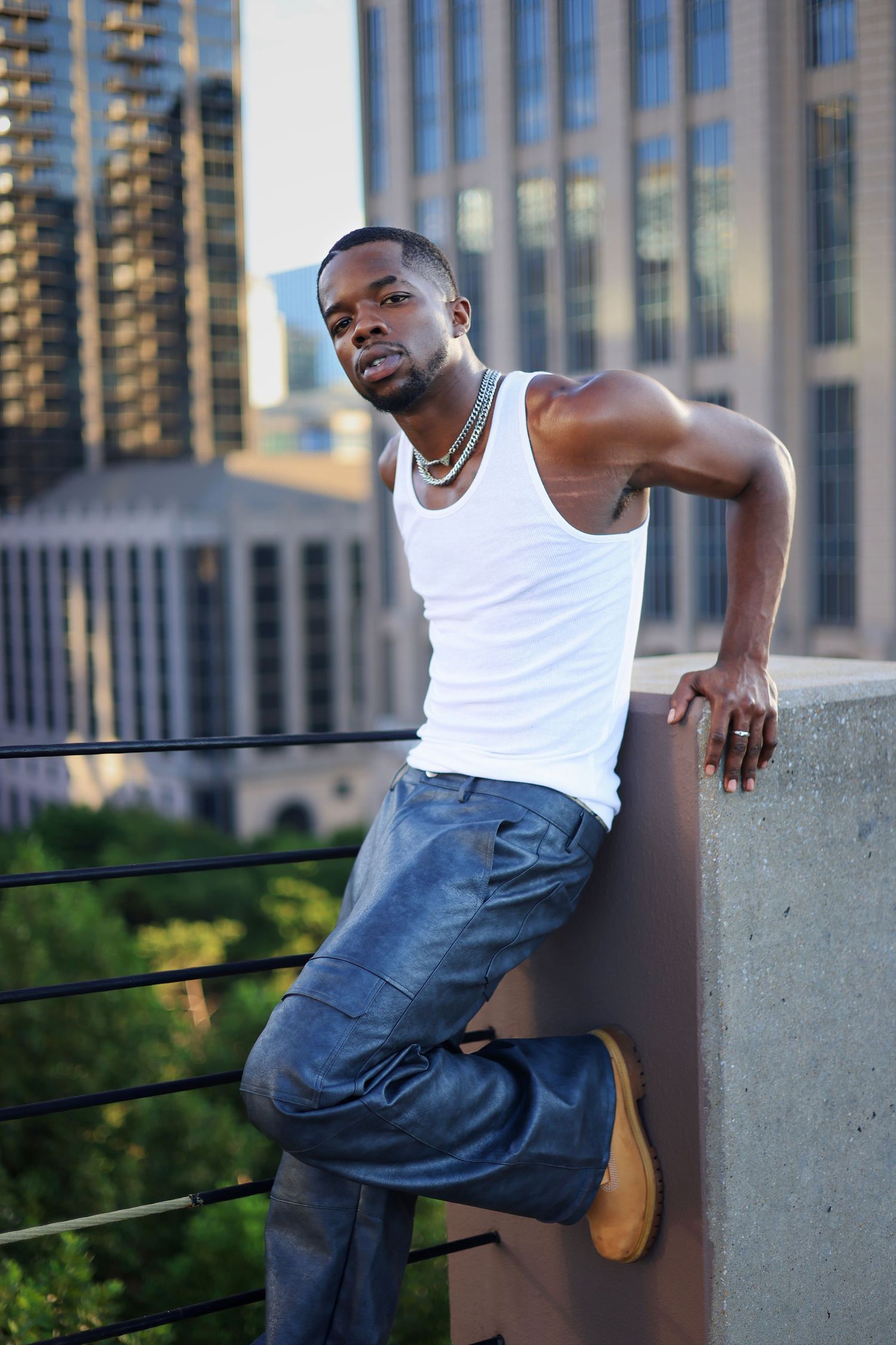 Fit man in white tank top leaning casually on rooftop ledge with city skyline backdrop.