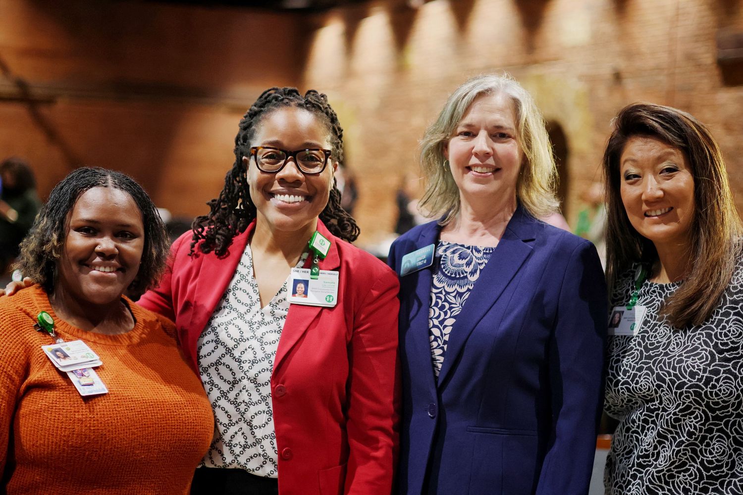 Four professional women smiling together at a formal indoor event, wearing business attire with name badges.