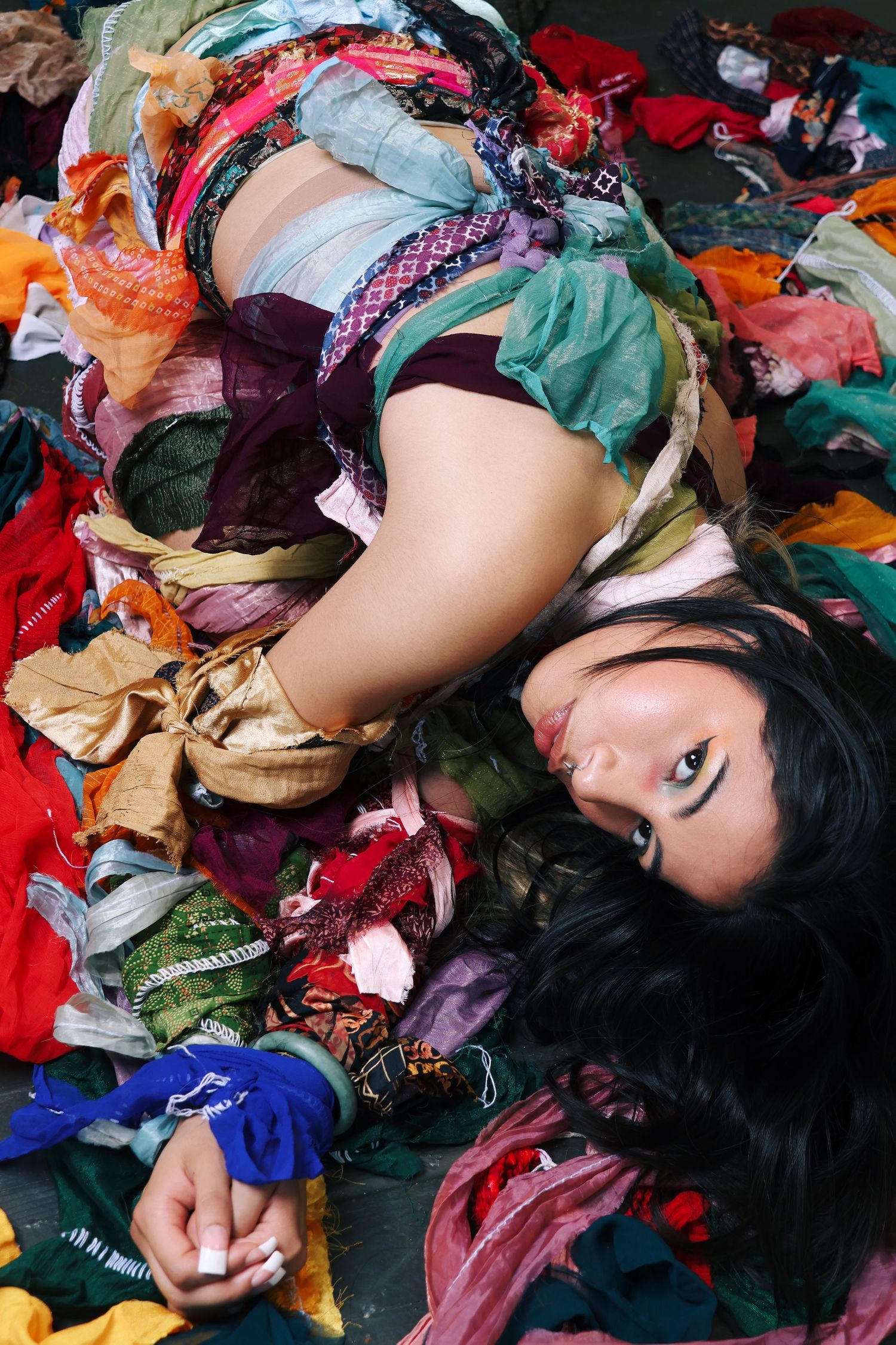 Woman lying on a colorful pile of fabric scraps and clothing, looking up at the camera with dark hair spread out.