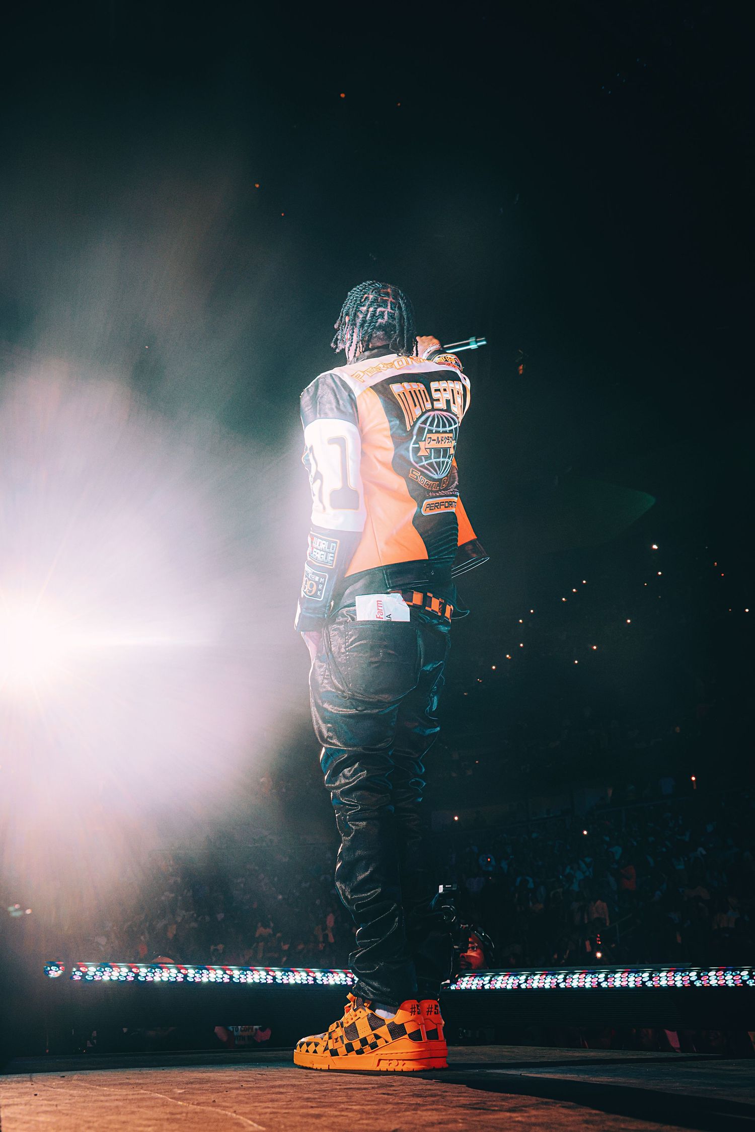 Rapper performing on stage in orange jersey and colorful sneakers, backlit by dramatic spotlight with smoke effects.
