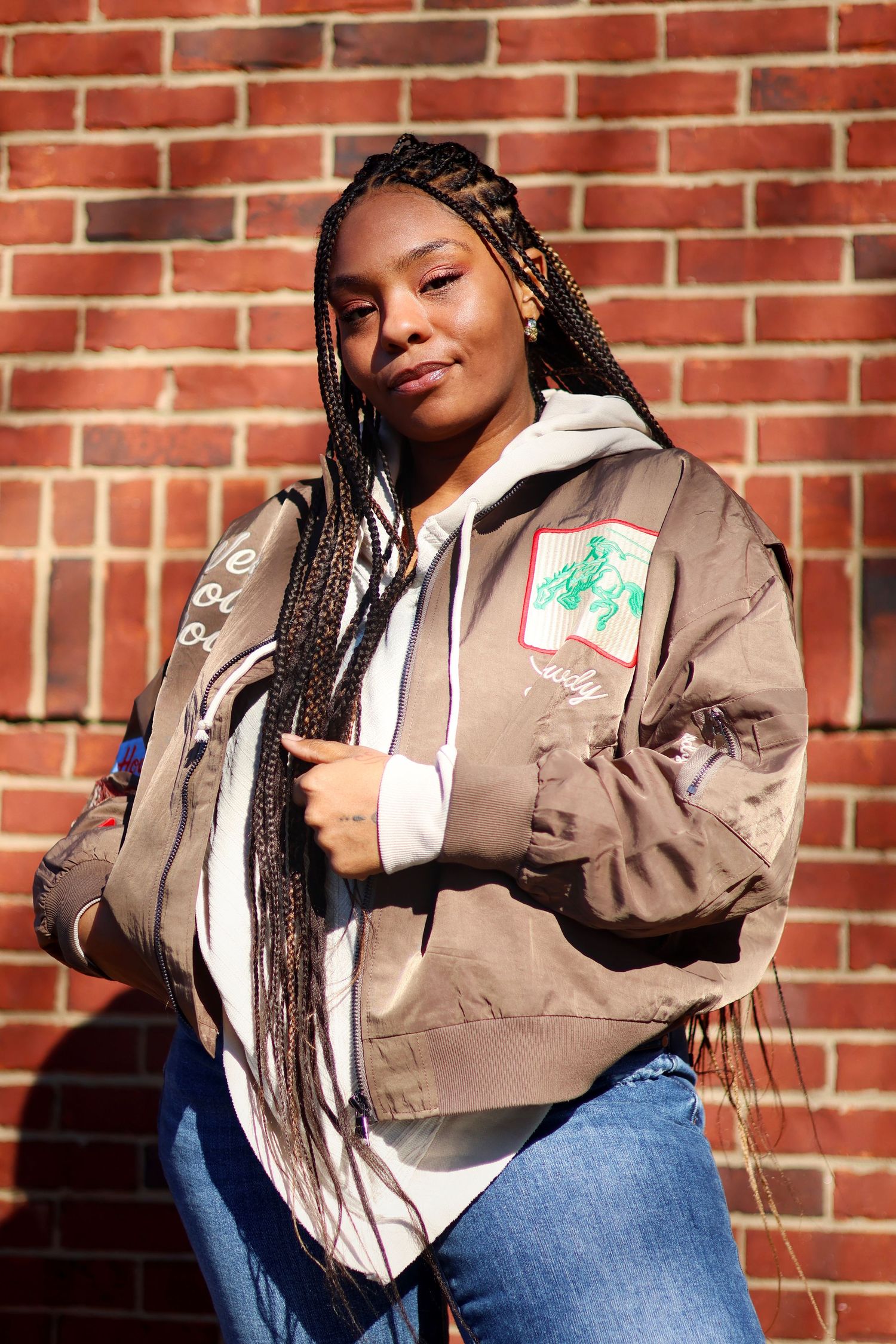 Woman with box braids wearing bomber jacket and hoodie posing against brick wall outdoors.