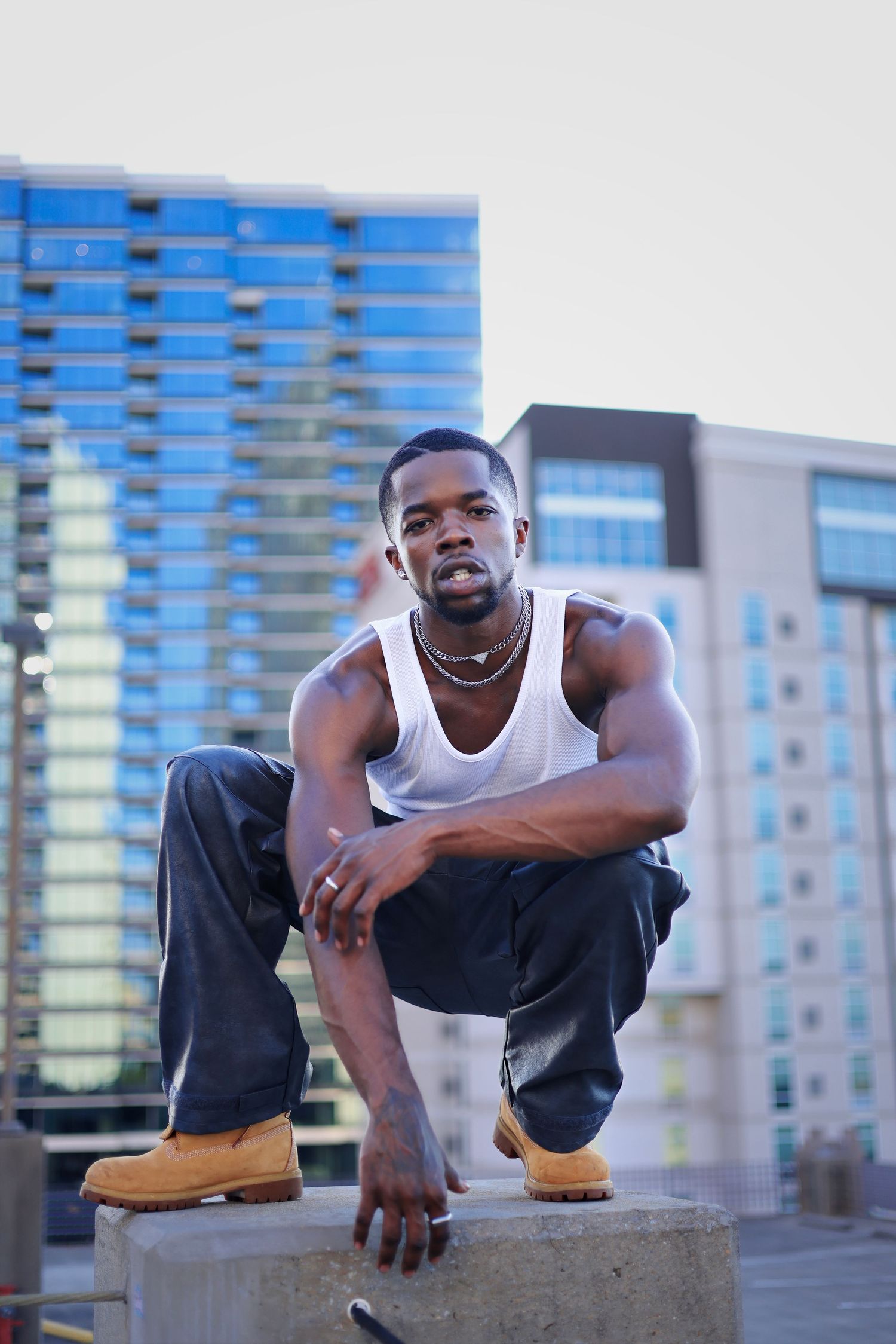 Athletic Black man in white tank and jeans crouching on urban rooftop with city skyline.