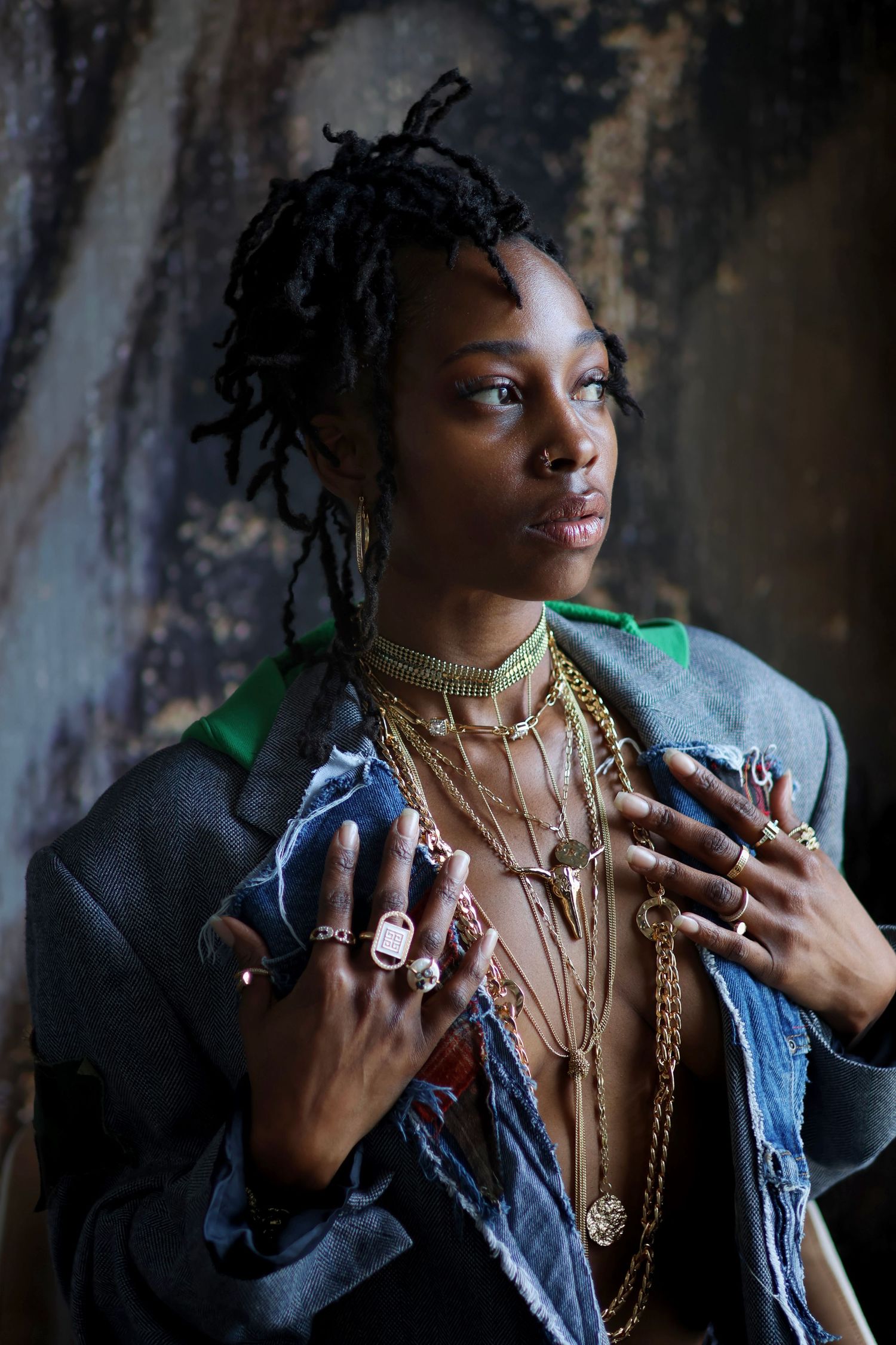 Woman with locs wearing layered gold necklaces and rings with denim jacket in editorial shot.