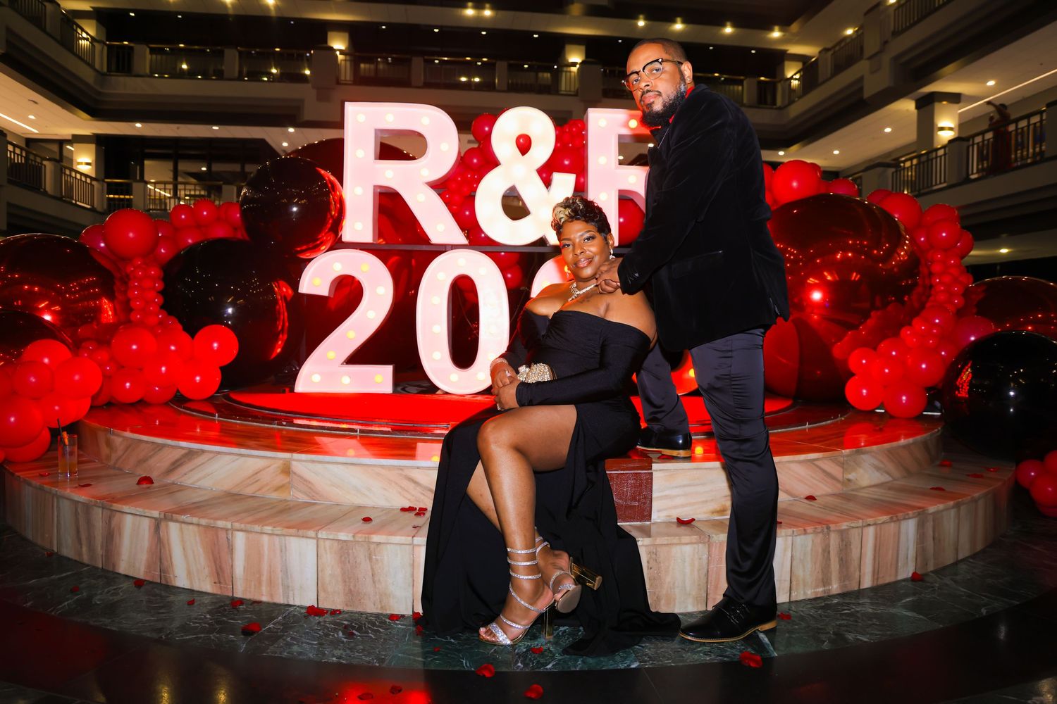 Couple posing elegantly in front of R&B 20 sign with red balloons at a glamorous event.