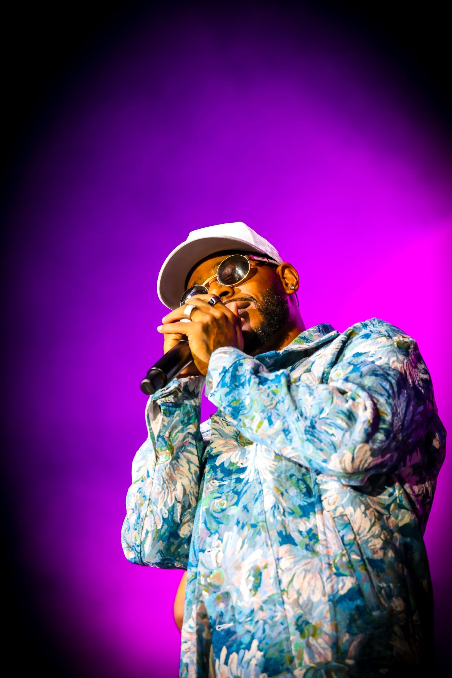 A male performer in a floral jacket and white cap sings into a microphone on a purple-lit stage.