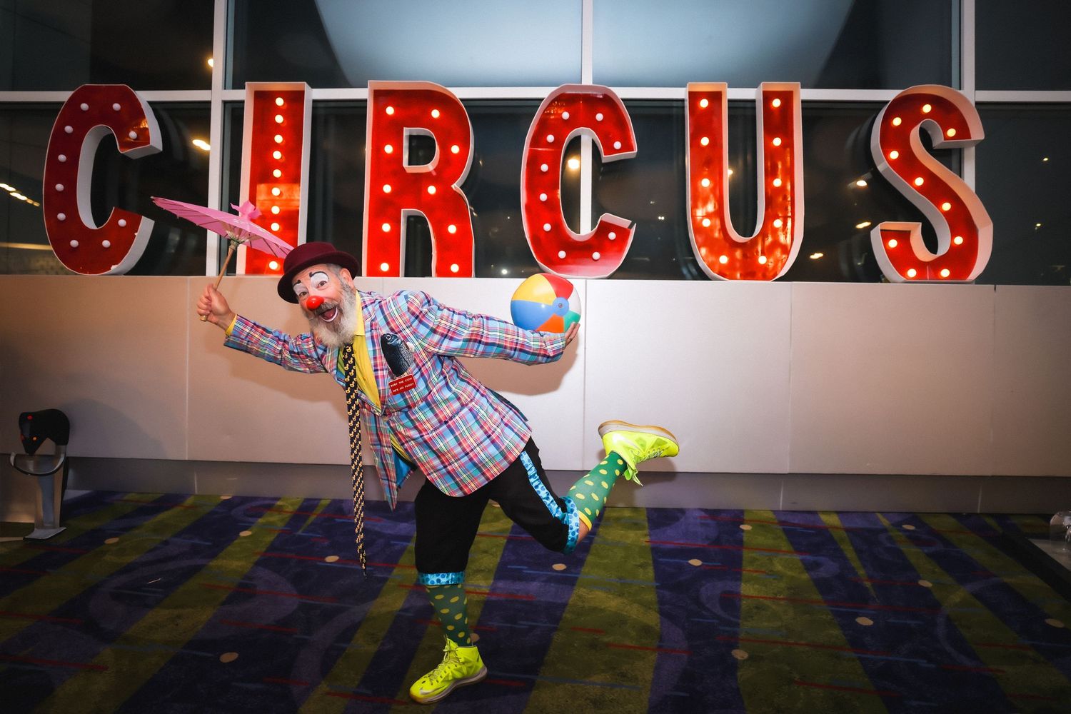 A colorful clown poses playfully with balloon animals in front of a large illuminated CIRCUS sign.