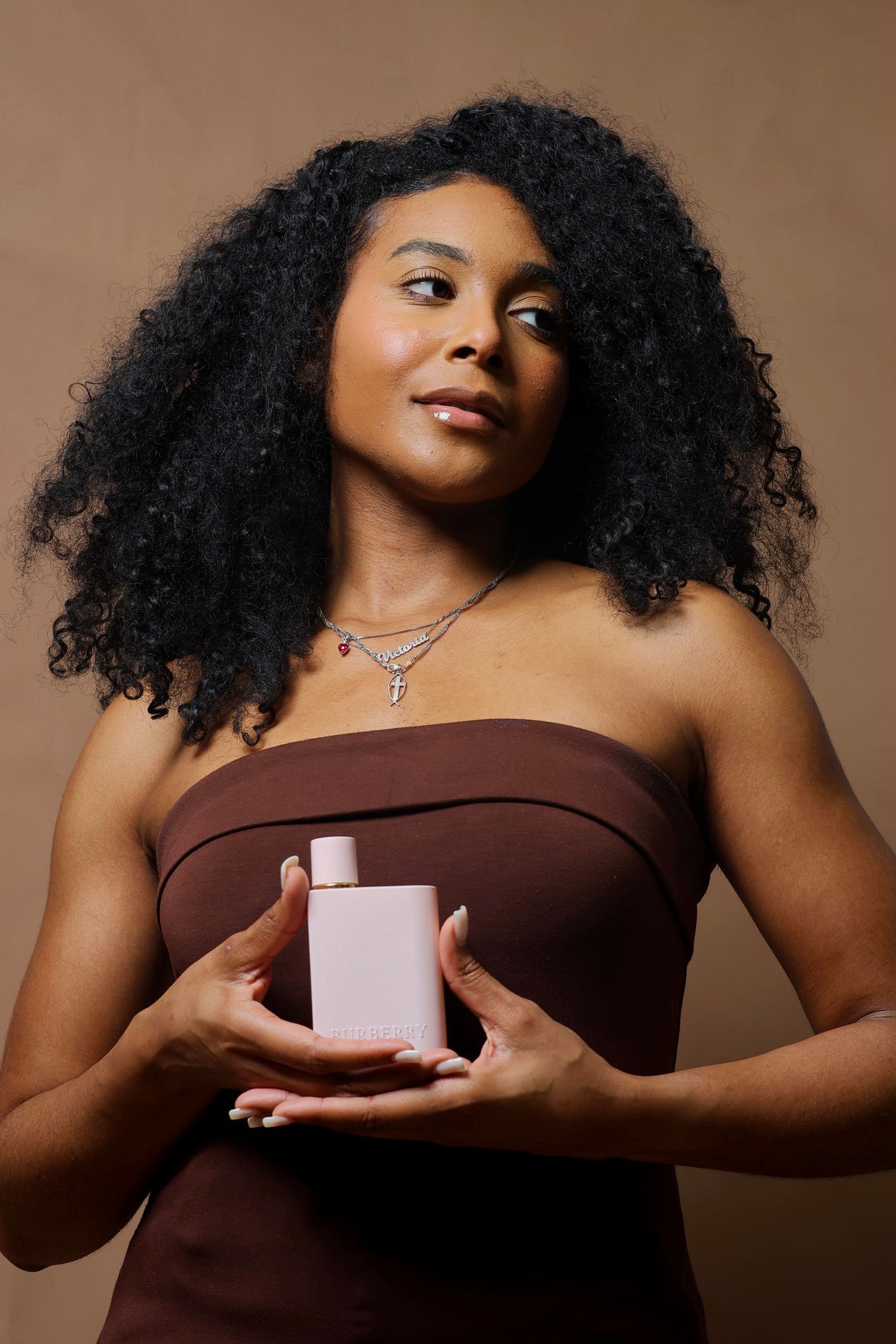 Woman with curly hair in brown strapless top holding a pink perfume bottle, wearing layered necklaces.