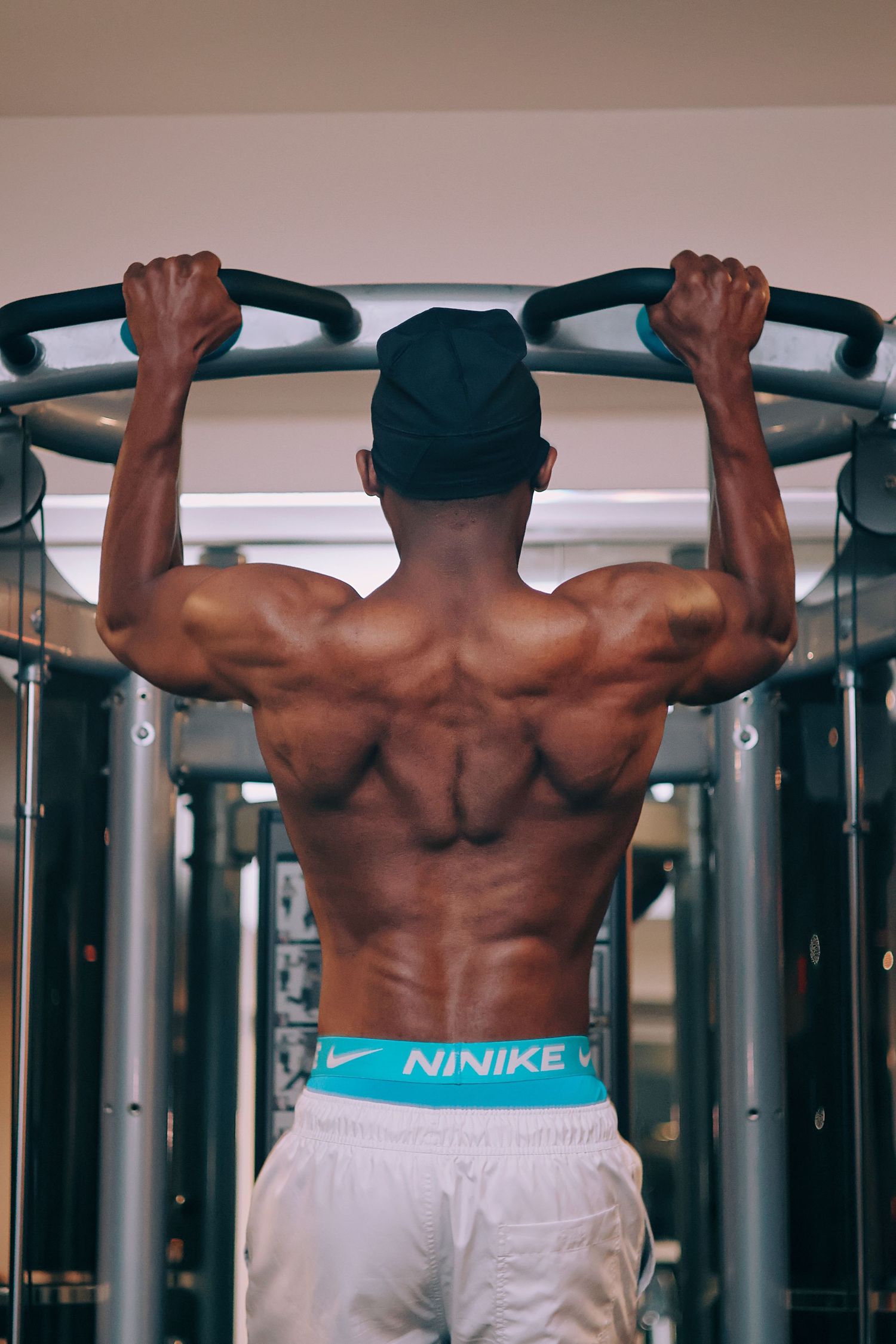 Muscular man doing pull-ups on a gym bar, showing defined back muscles, wearing Nike waistband and white shorts.