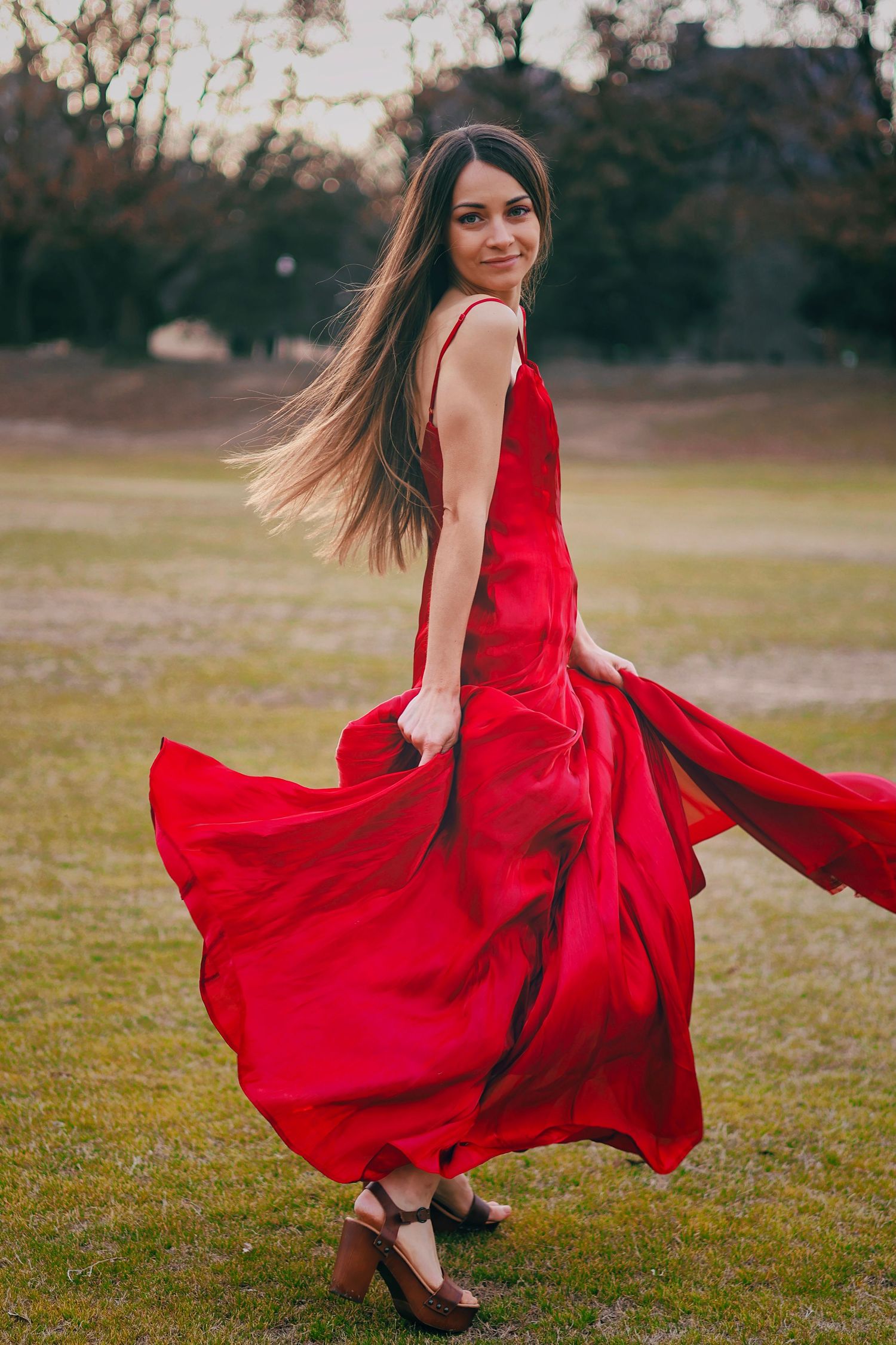 Young woman twirling in elegant red maxi dress outdoors in park.