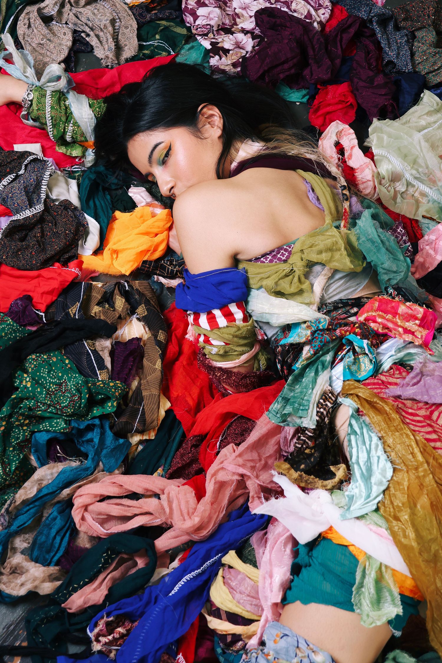 Young woman lying among a colorful pile of scattered clothing and fabric on the floor.